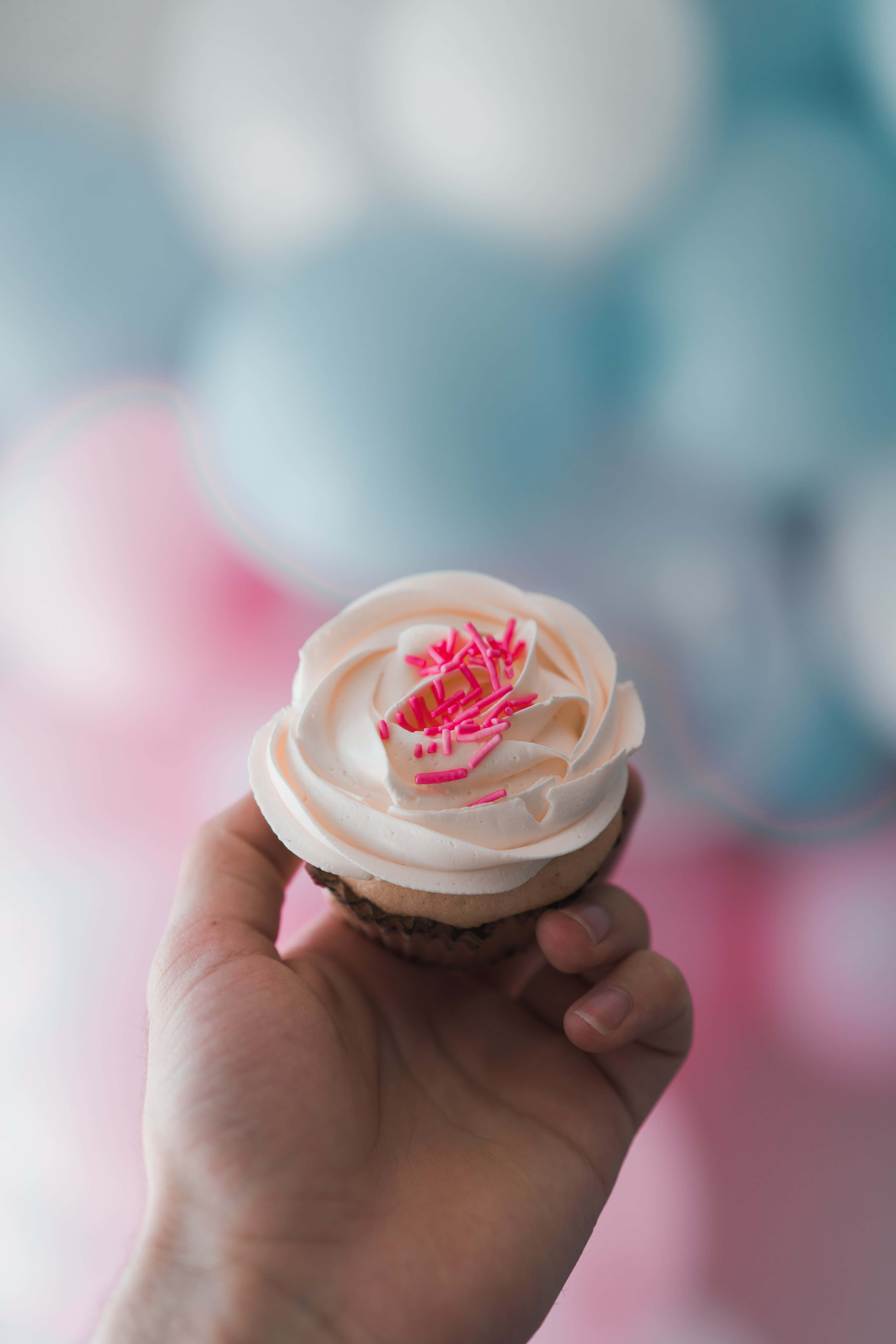Cupcake 桌布 壁紙 — 一隻手拿著一個有粉紅色糖霜的杯子蛋糕，背景是柔和模糊的粉彩氣球。