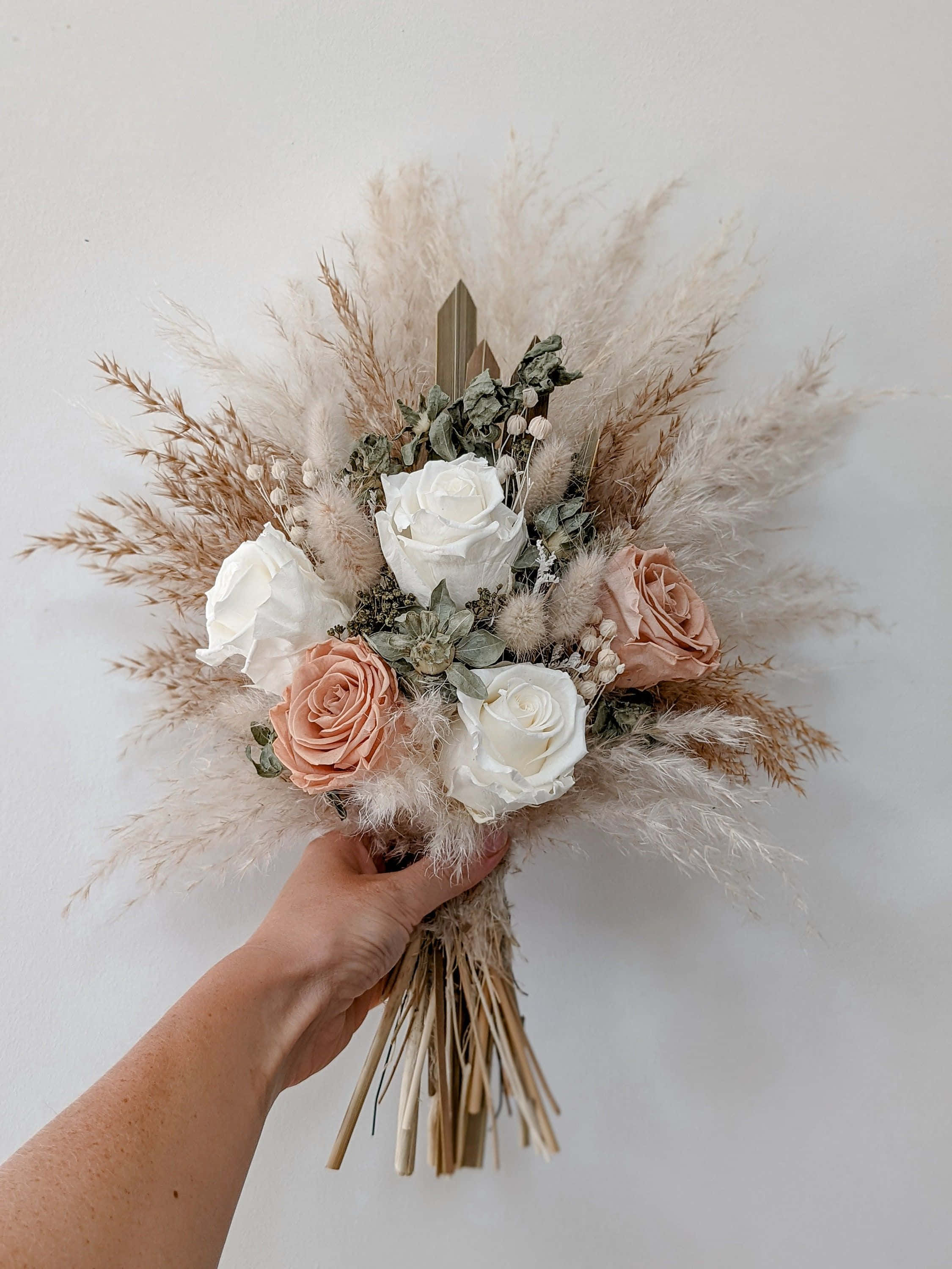 Roses Wedding 桌布 壁紙 — 一隻手拿著一個柔軟的波希米亞風格花束，包含白色和蜜桃色玫瑰以及羽毛狀的蒲葦，背景是純白牆壁。