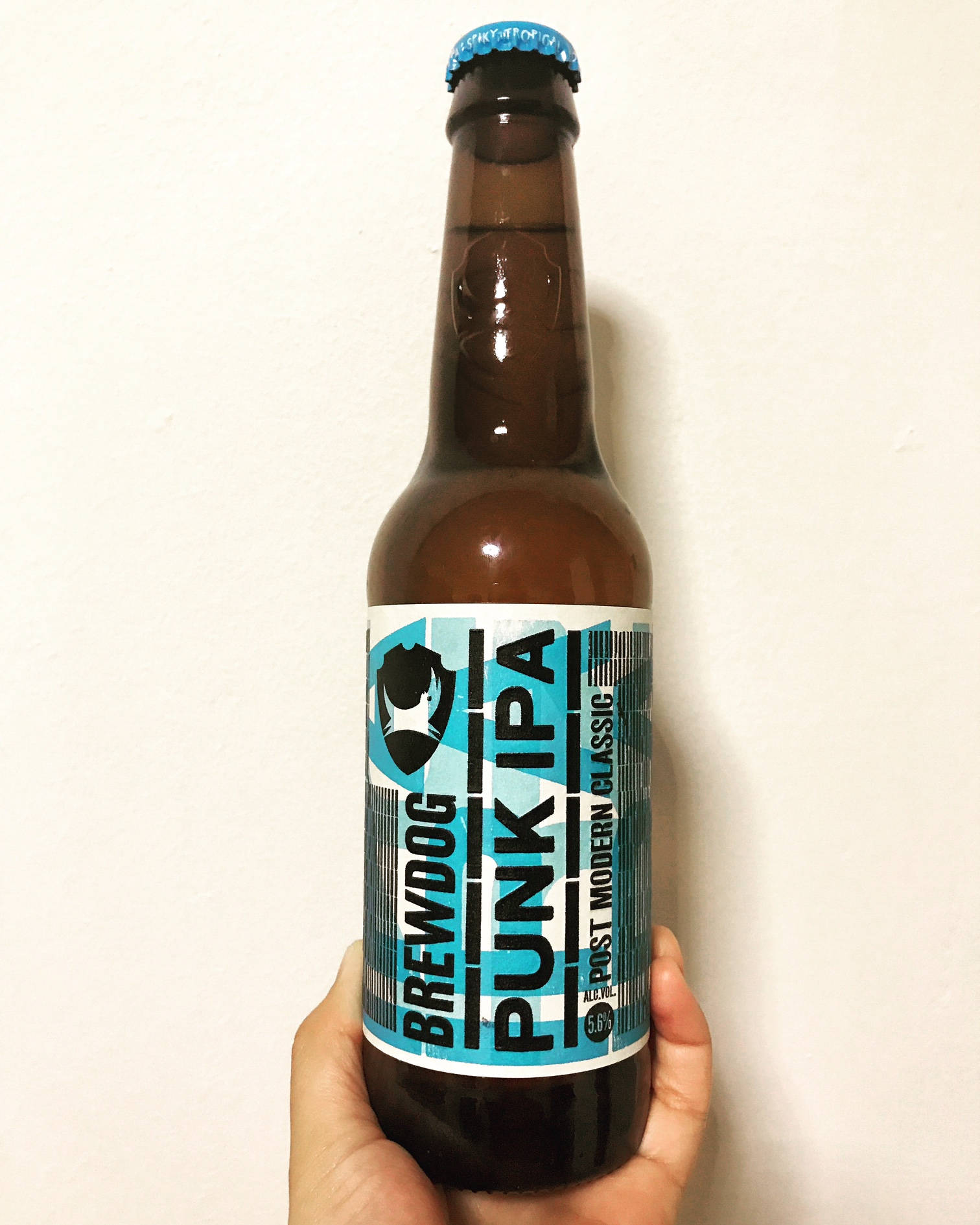 Drink 桌布 壁紙 — 一隻手拿著一個棕色 BrewDog Punk IPA 啤酒瓶，對著白色素牆，瓶蓋為藍色，標籤大膽。