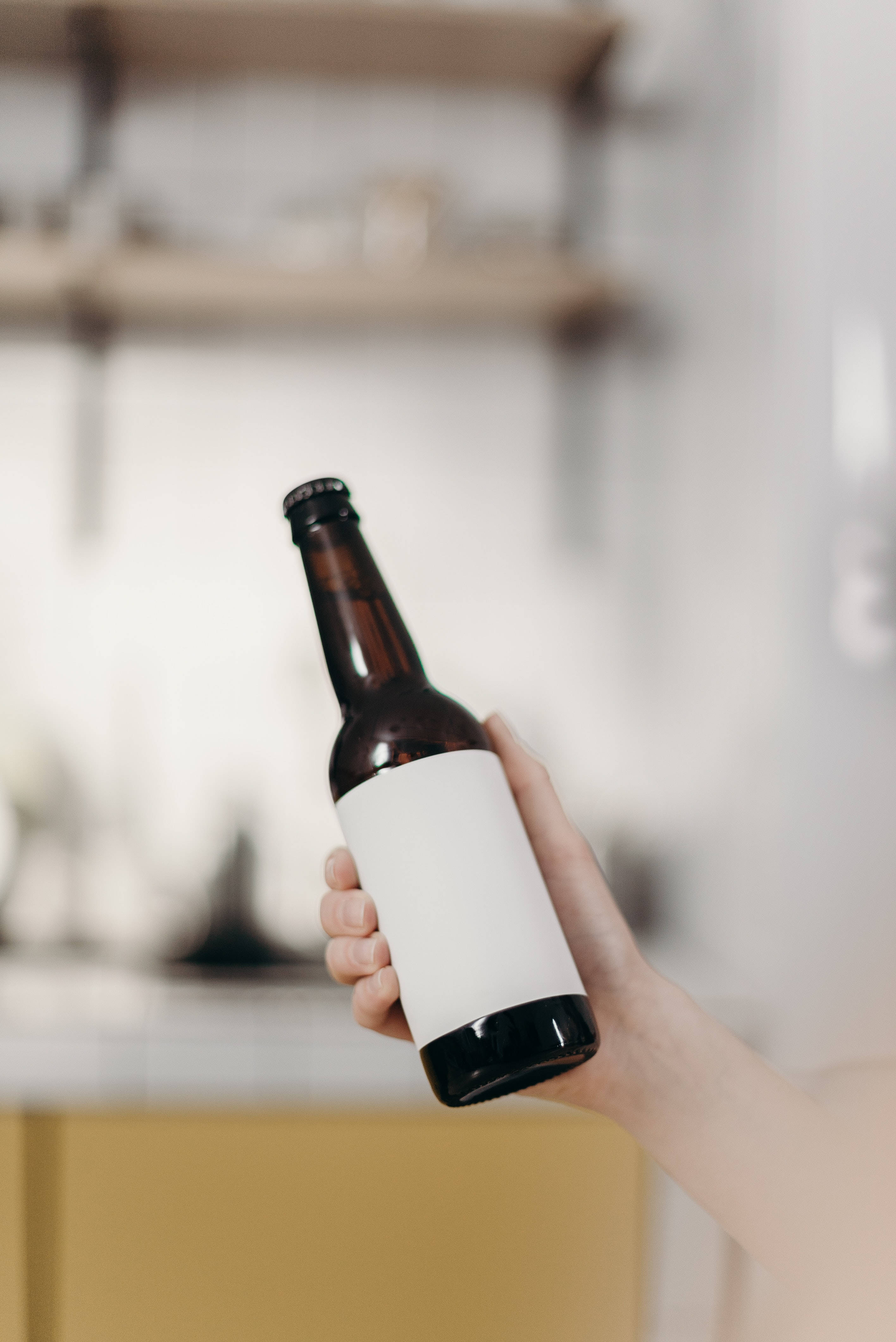 alcohol 桌布 壁紙 — 一隻手拿著一個棕色啤酒瓶，帶有空白白色標籤，背景是柔和模糊的廚房。
