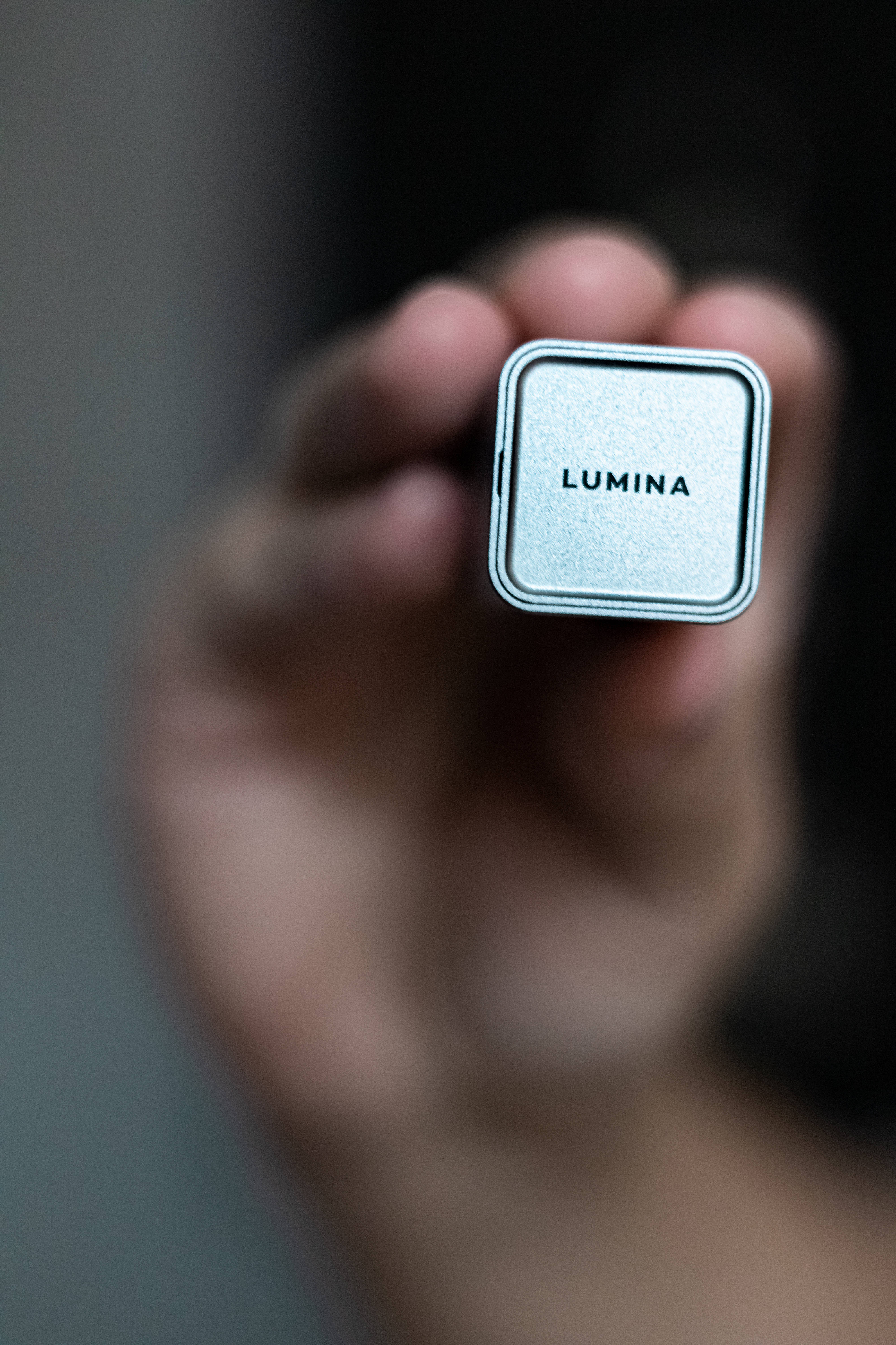 Webcam 桌布 壁紙 — 一隻手拿著一個標有「LUMINA」的小型銀色方形裝置，背景為模糊的灰色。