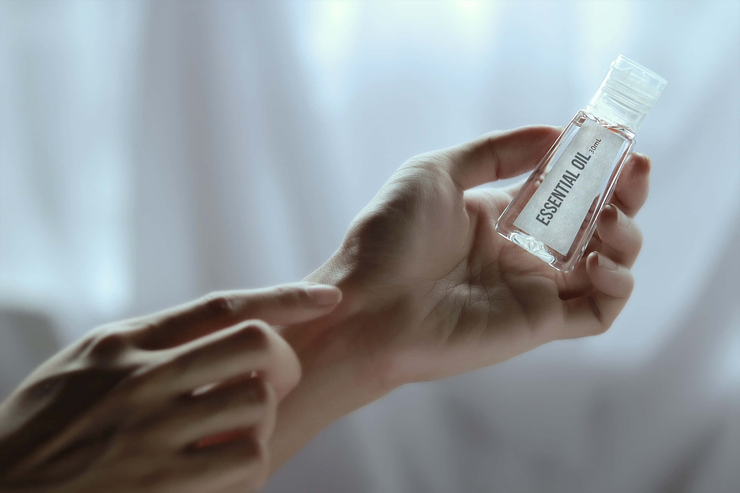 hands 桌布 壁紙 — 一隻手拿著一個標籤為精油的透明瓶子，另一隻手輕輕地將幾滴精油滴在手腕上，背景是柔軟的白色窗簾。