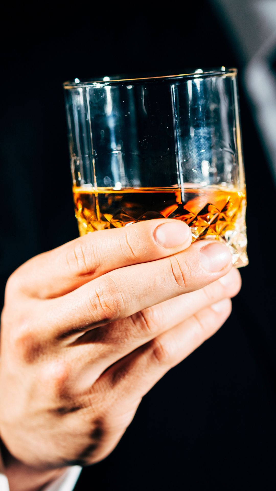 drinks 桌布 壁紙 — 一隻手拿著一個水晶杯，裡面裝著琥珀色的威士忌，背景昏暗，光線柔和。