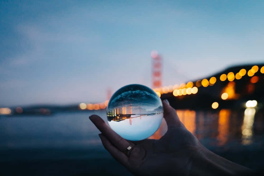 reflection 桌布 壁紙 — 一隻手拿著一個水晶球，反射出暮色中的金門大橋，背景是模糊的城市燈光。