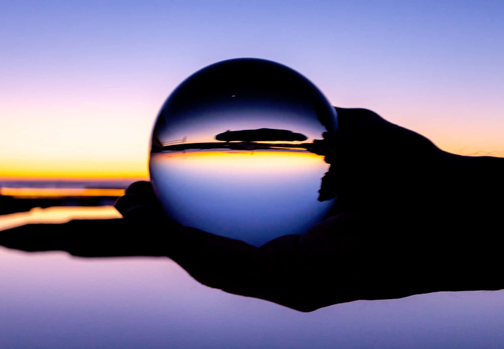 reflection 桌布 壁紙 — 一隻手拿著一個水晶球，反射出水上充滿活力的日落，遠處有一艘船的剪影。