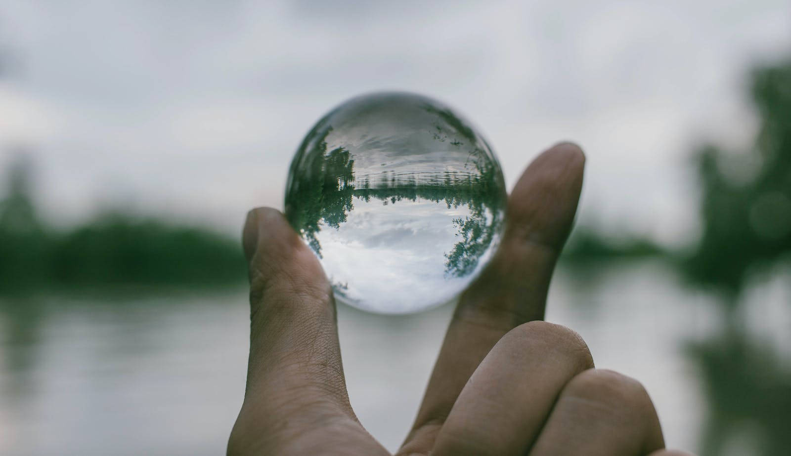 Transparent 桌布 壁紙 — 一隻手拿著一個水晶球，反射出陰天下一片寧靜的湖泊和樹木。
