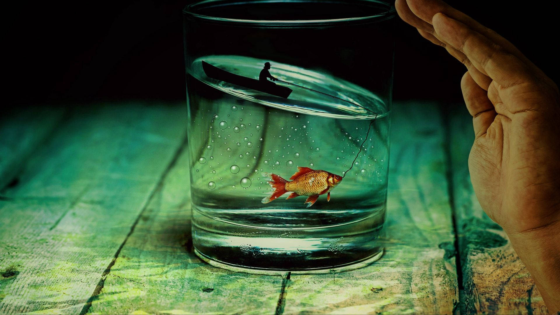 釣魚 桌布 壁紙 — 一隻手拿著一個玻璃杯，裡面有一條金魚在釣魚線上，上方有一個小船和人物在綠色木質表面上。