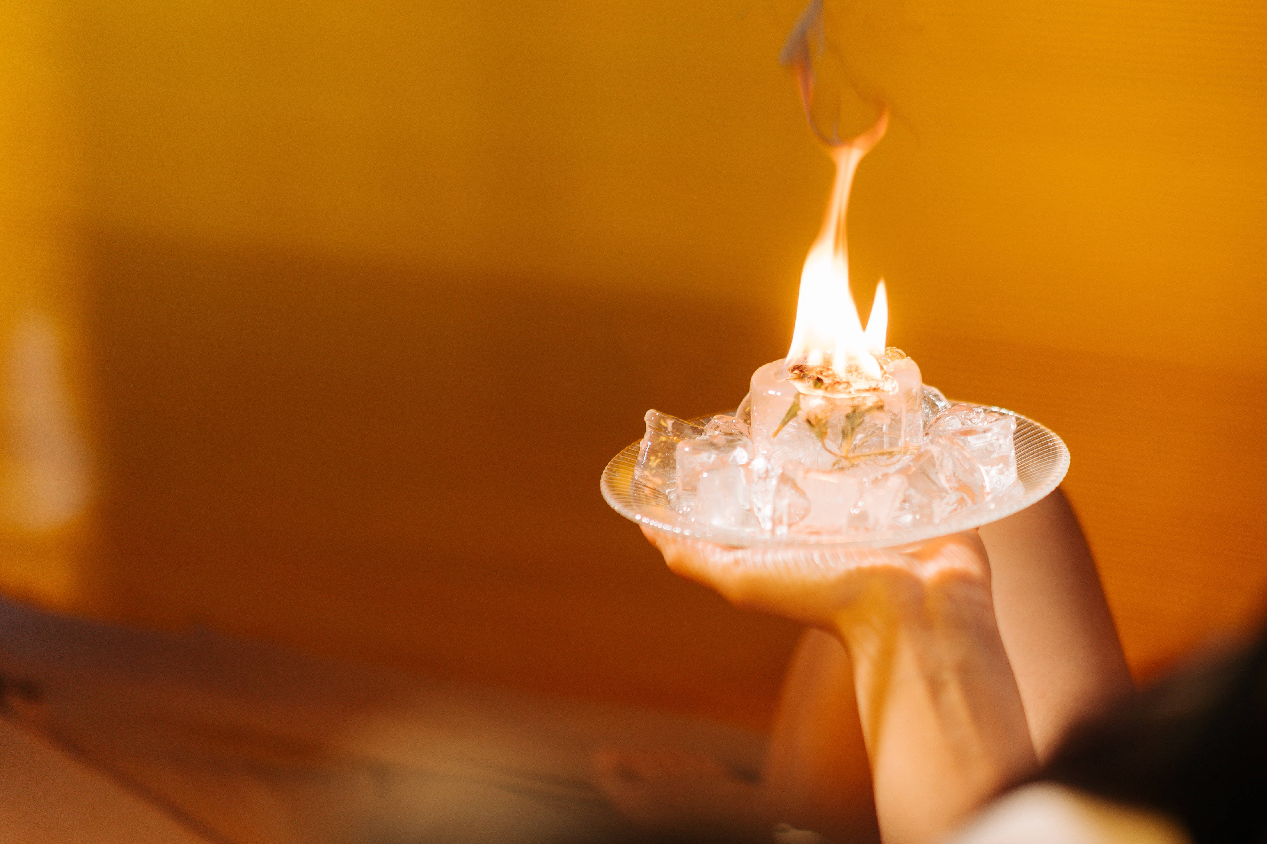 Fire And Ice 桌布 壁紙 — 一隻手拿著一個玻璃碟，裡面有冰塊和一小團火焰從中升起，沐浴在溫暖的金色光芒中。