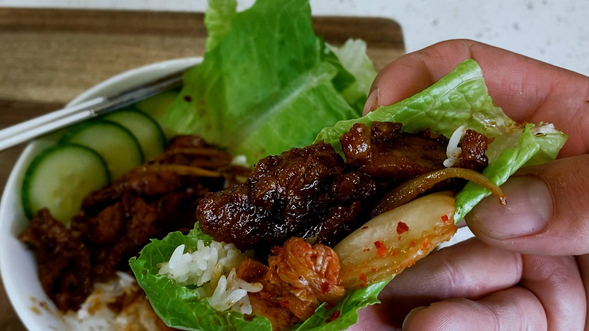 肉 桌布 壁紙 — 一隻手拿著一個生菜包，裡面有照燒肉、泡菜和米飯，放在一碗米飯、小黃瓜和蔬菜上面。
