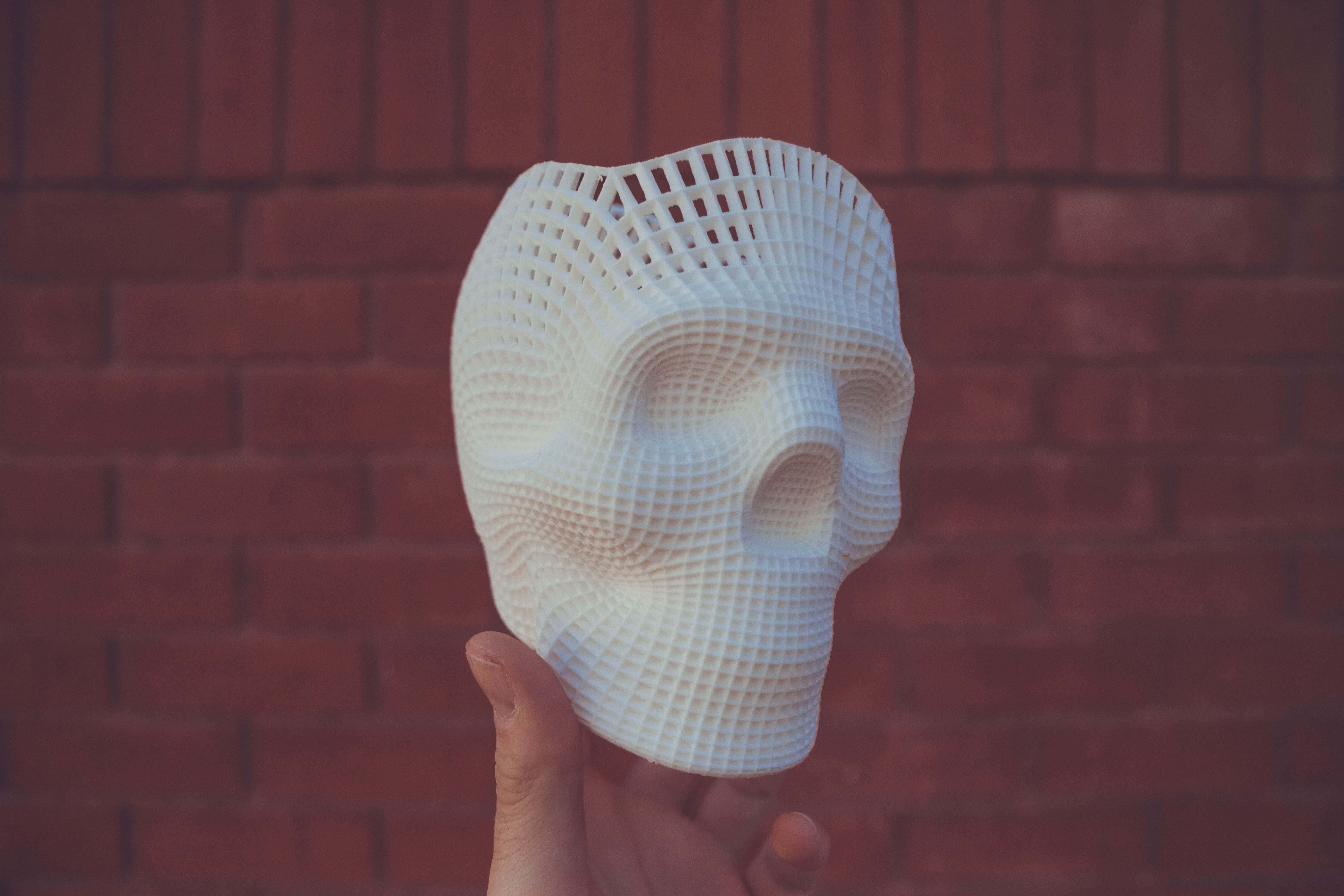 攝影 桌布 壁紙 — 一隻手拿著一個白色3D列印的網格圖案頭骨，背景是紅磚牆。