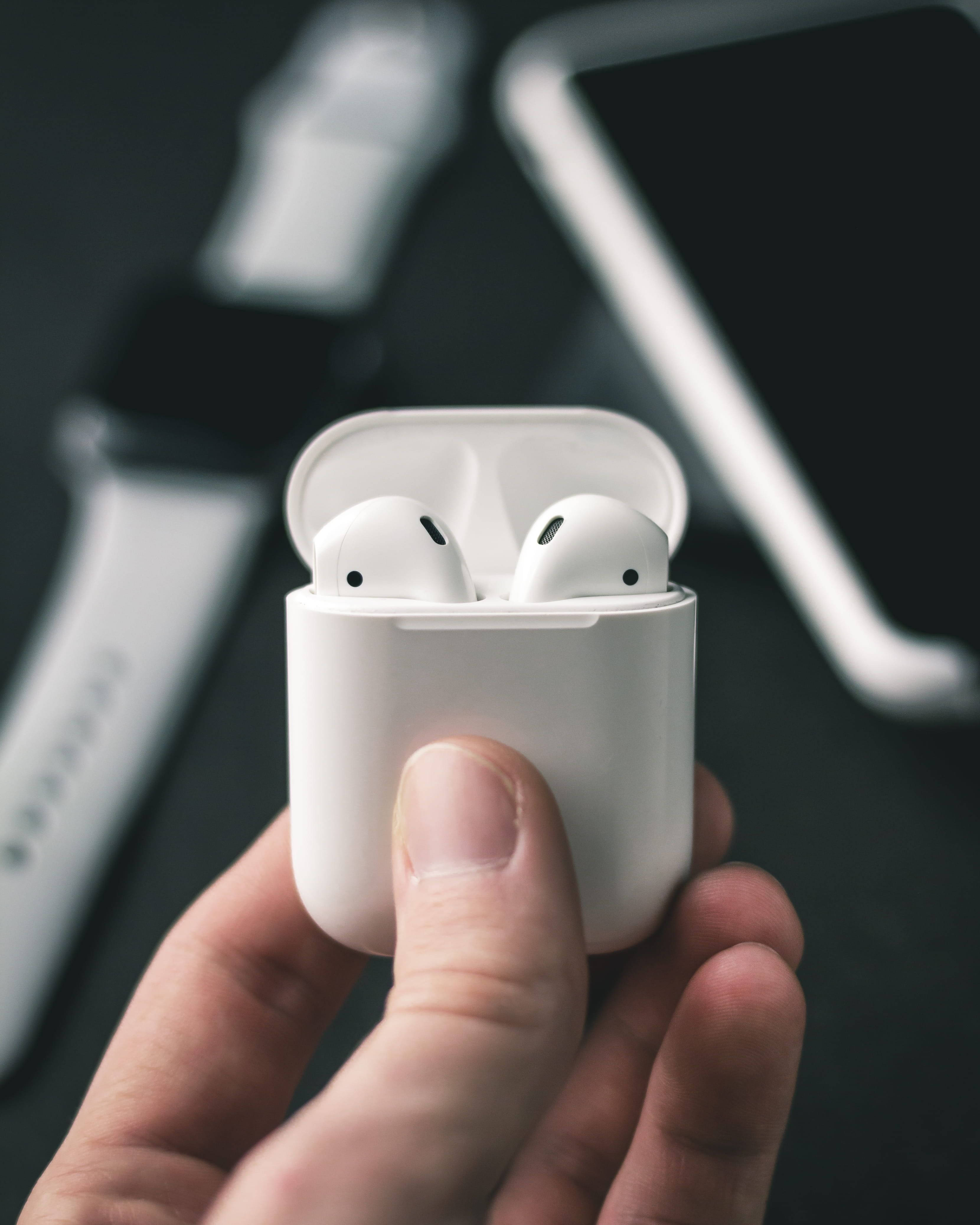Airpods 桌布 壁紙 — 一隻手拿著一個白色打開的AirPods充電盒，露出耳機，背景有模糊的智慧手錶和手機。