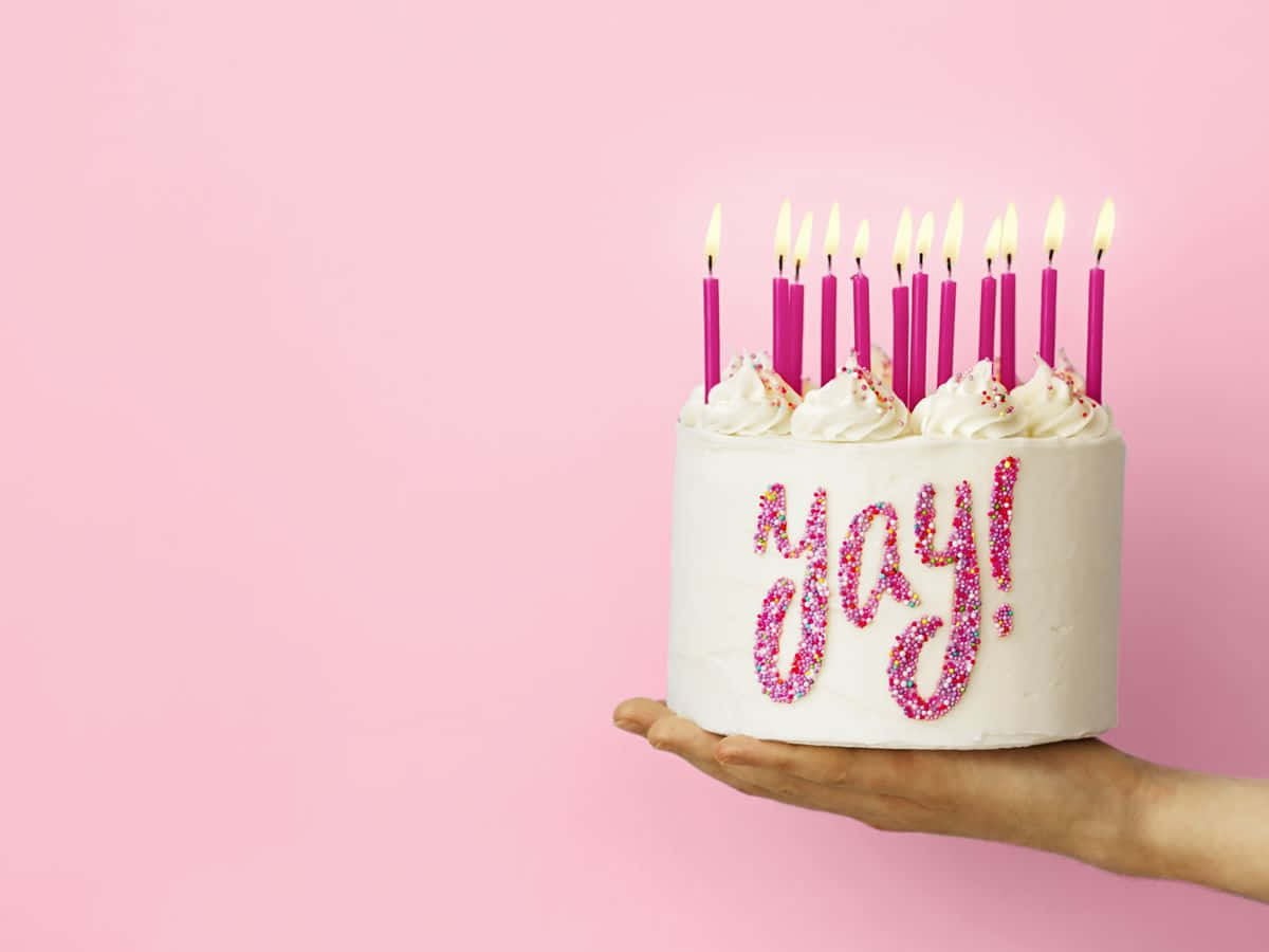 Birthday Cake 桌布 壁紙 — 一隻手拿著一個白色生日蛋糕，上面有粉紅色蠟燭和閃亮的yay字樣，背景是柔和的粉紅色。