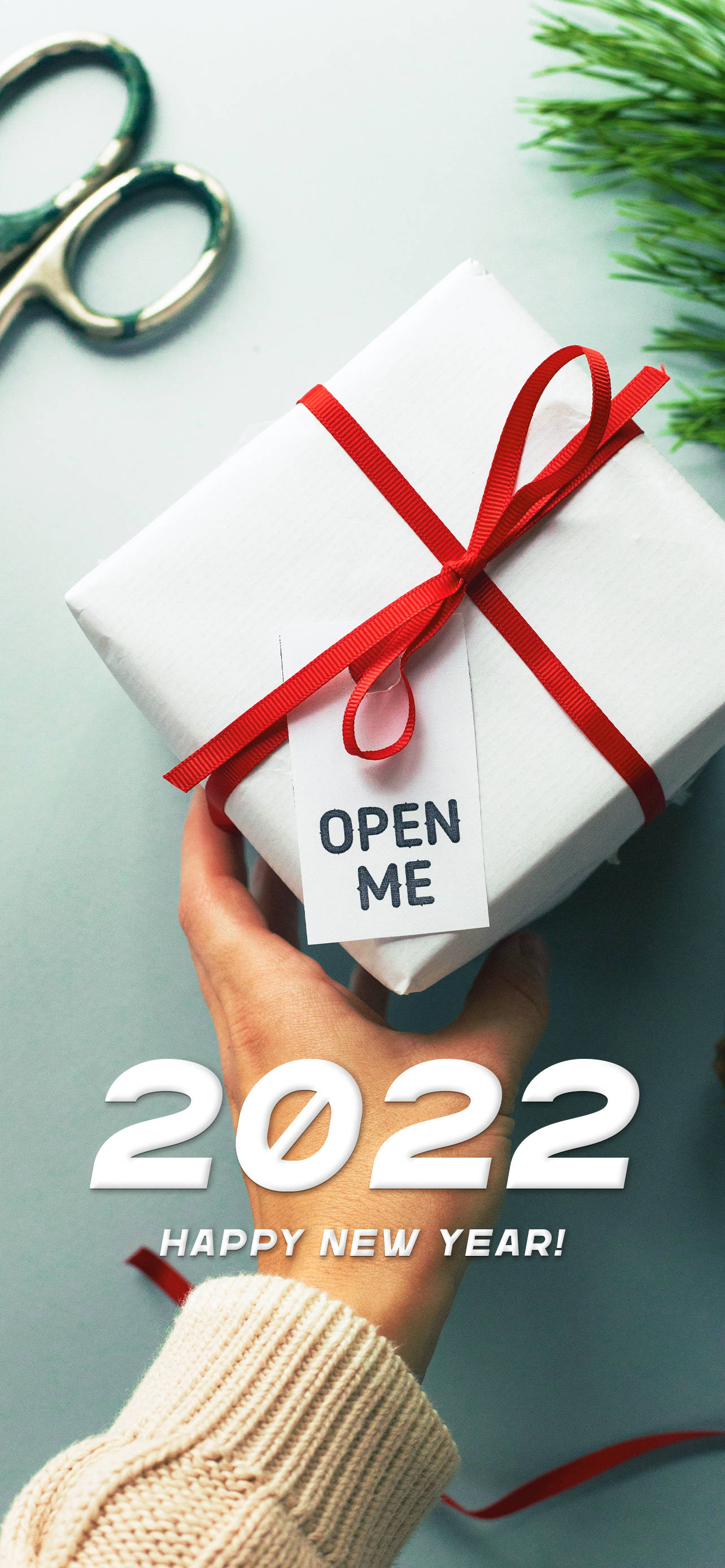 Happy New Year 2022 桌布 壁紙 — 一隻手拿著一個白色禮物盒，繫著紅色絲帶和一個寫著「打開我」的標籤，背景是藍綠色，有松針和剪刀。