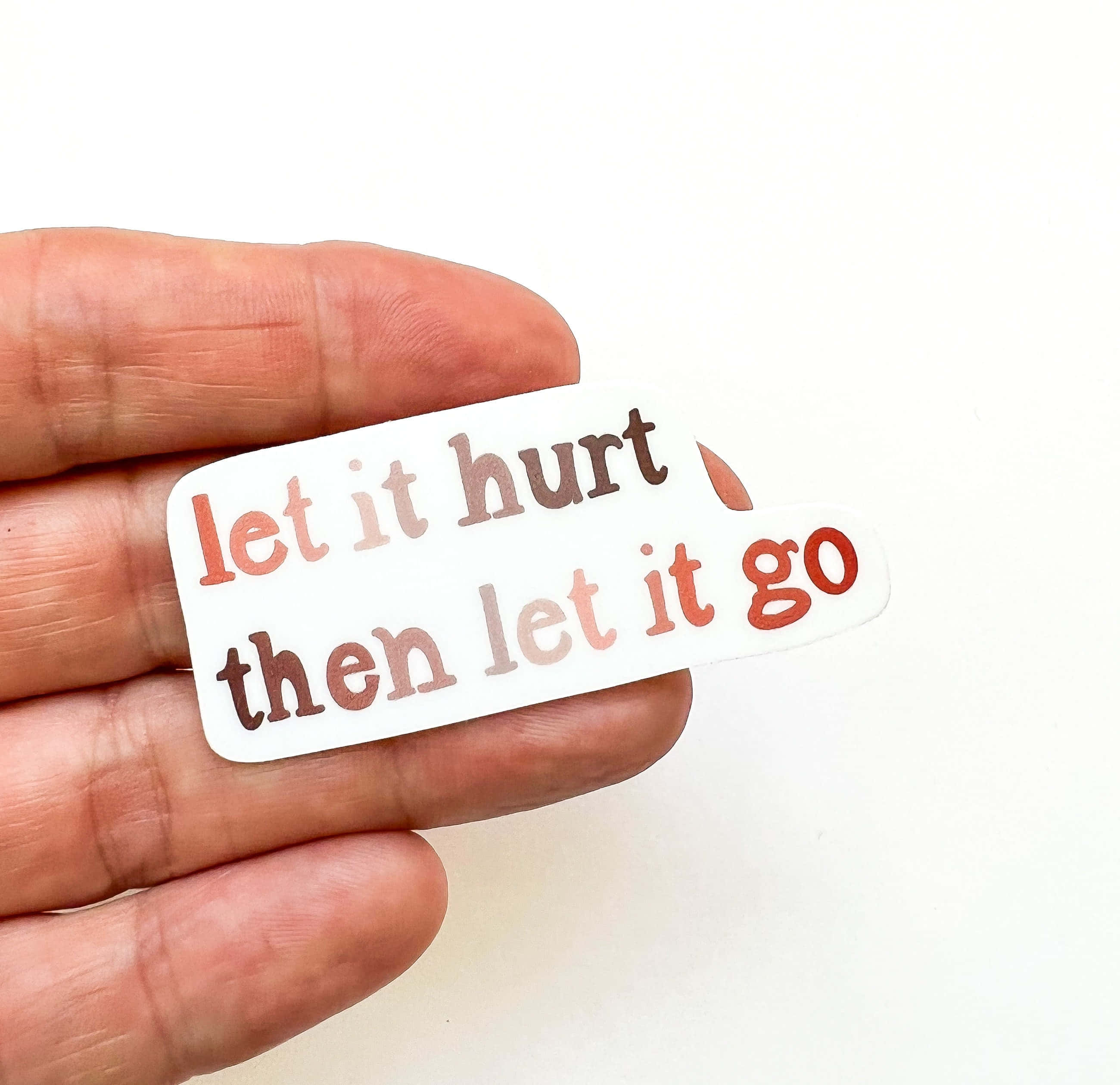 let it go 桌布 壁紙 — 一隻手拿著一個白色貼紙，上面有漸層橙色和棕色的文字「let it hurt then let it go」。