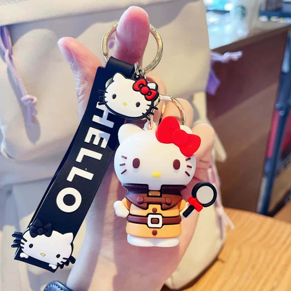 Kawaii Hello Kitty 桌布 壁紙 — 一隻手拿著一個穿著偵探裝、手持放大鏡的Hello Kitty鑰匙圈，背景是模糊的室內環境。