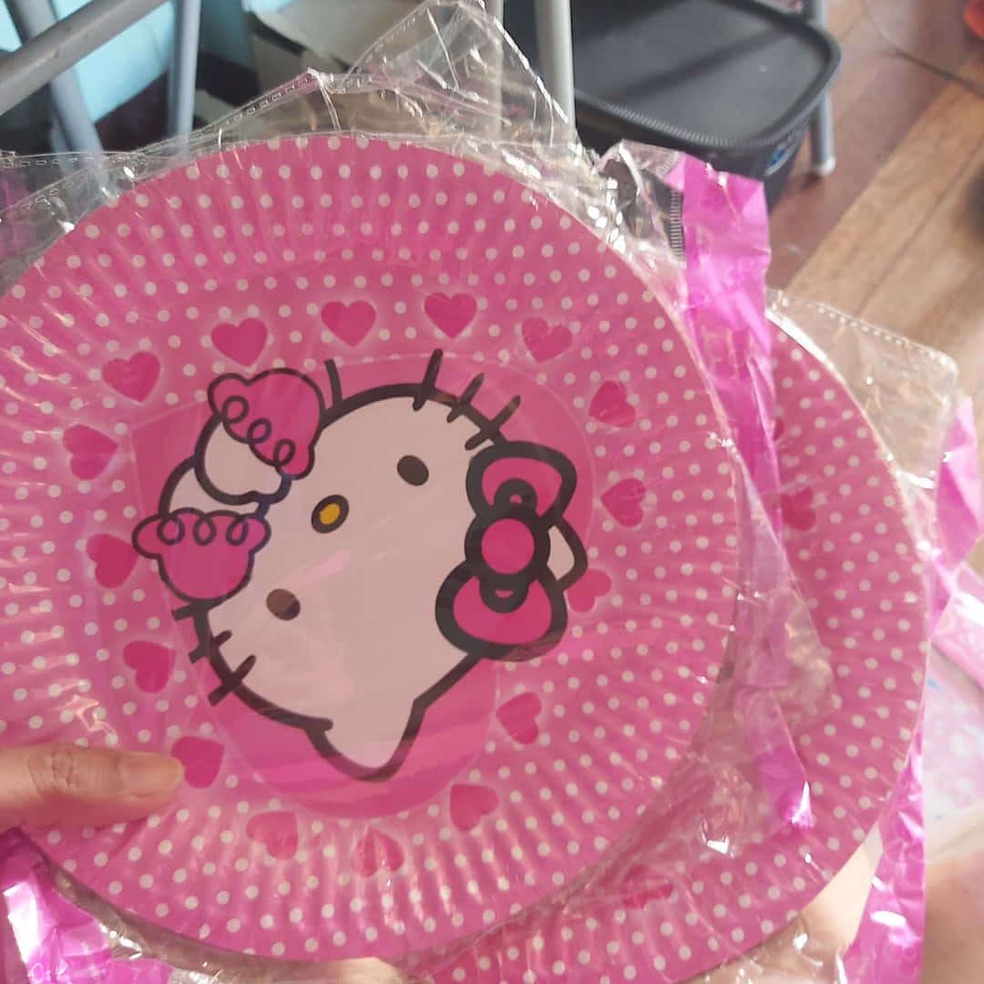 Hello Kitty Party 桌布 壁紙 — 一隻手拿著一個粉紅色Hello Kitty紙盤，上面有白色圓點和愛心圖案，並用透明塑膠包裝著。