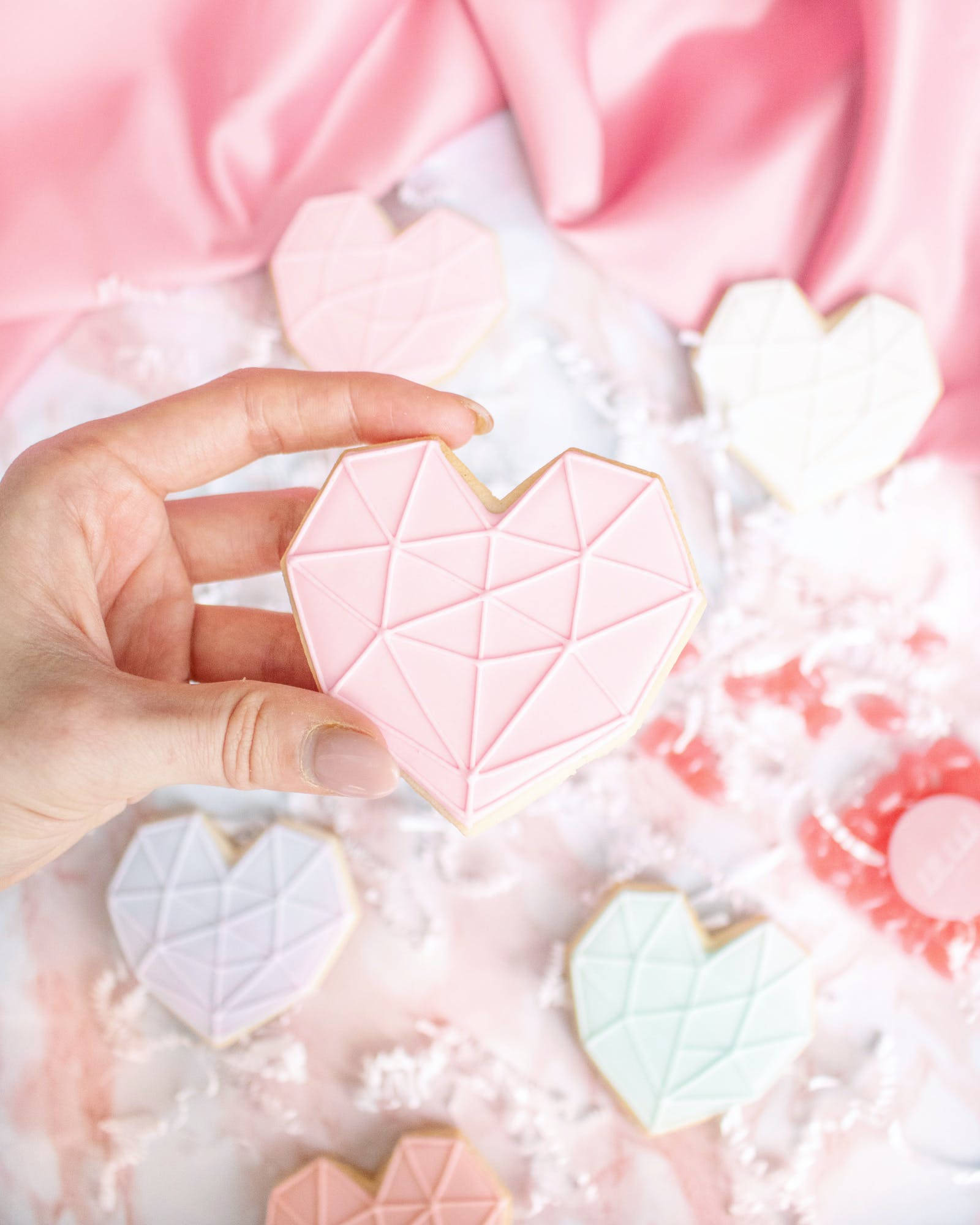 Pastel Pink Heart 桌布 壁紙 — 一隻手拿著一個粉紅色幾何心形餅乾，放在柔軟粉紅色布料和碎紙上的粉彩色心形餅乾上方。