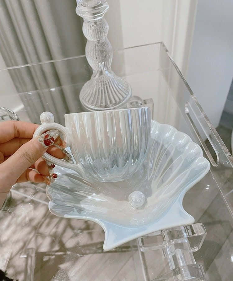 Teatime 桌布 壁紙 — 一隻手拿著一個精緻的棱紋玻璃杯，放在一個扇貝形白色碟子上，置於透明壓克力桌上，背景有一個玻璃燭台。
