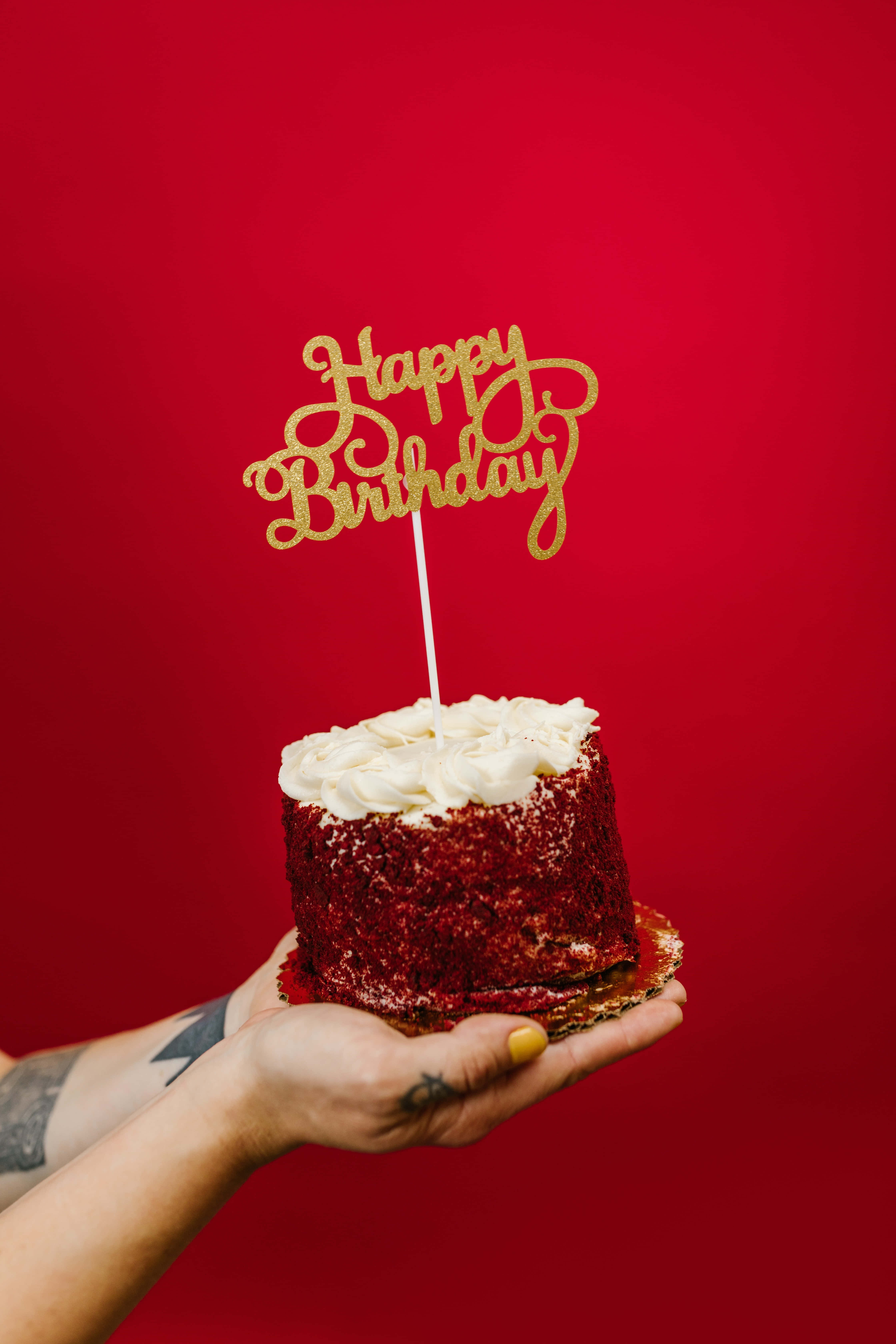 Cute Birthday 桌布 壁紙 — 一隻手拿著一個紅絲絨蛋糕，上面有白色糖霜和金色「Happy Birthday」裝飾，背景為純紅色。