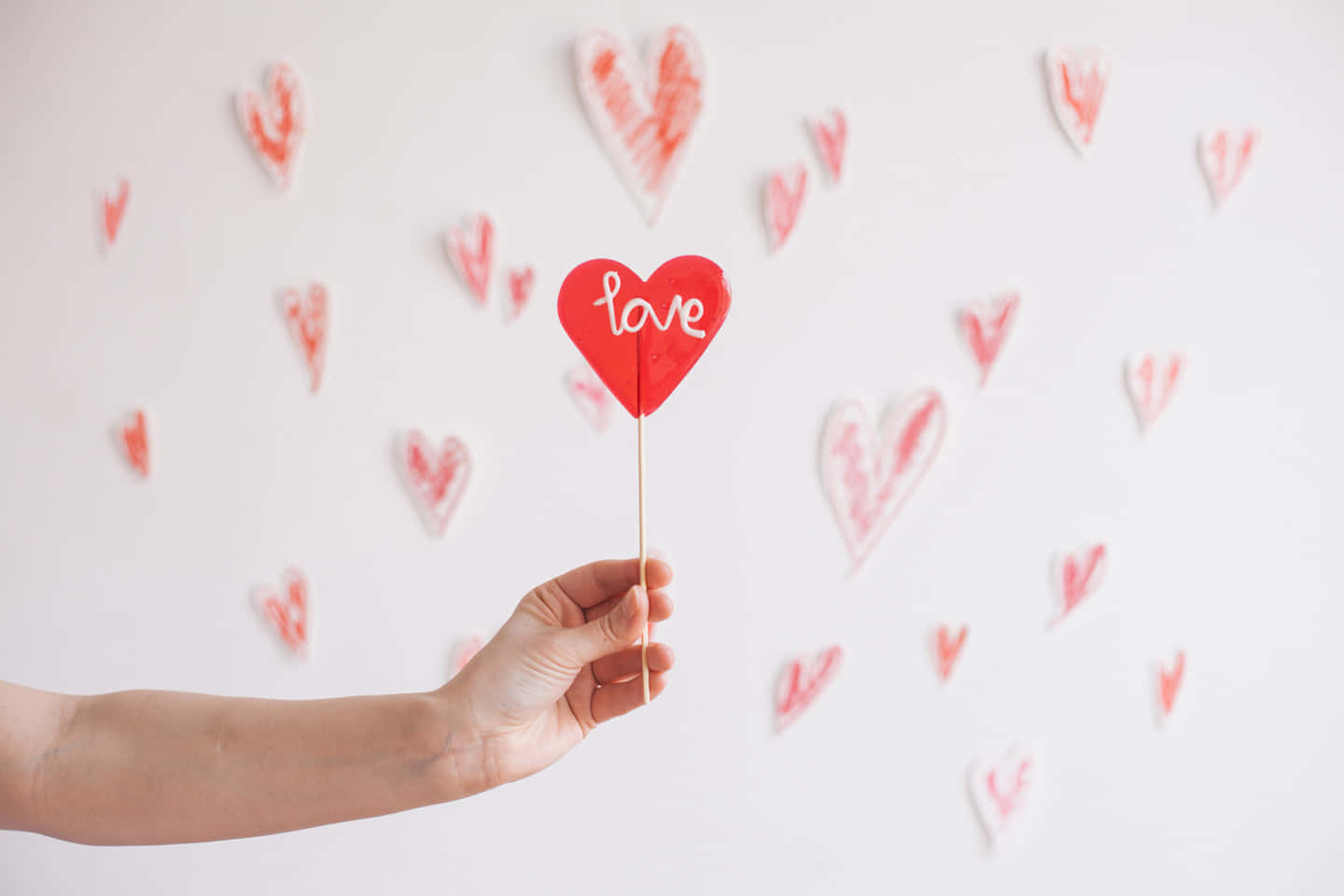 愛心 桌布 壁紙 — 一隻手拿著一個紅色心形標牌，上面寫著 love，對著一面裝飾著散落紅色心形的白色牆壁。