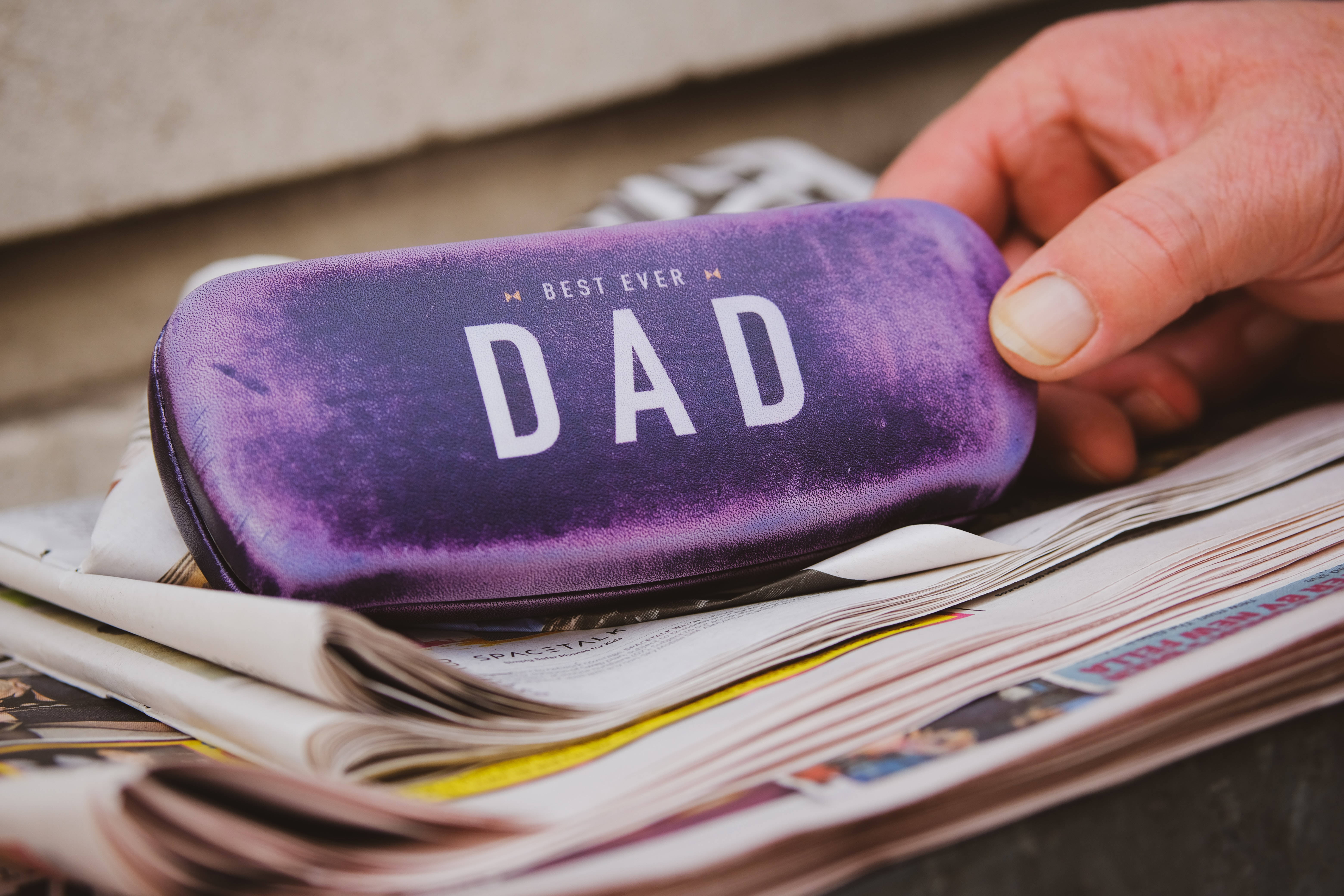 Dad 桌布 壁紙 — 一隻手拿著一個紫色眼鏡盒，上面有 'BEST EVER DAD' 字樣，放在一堆報紙上。