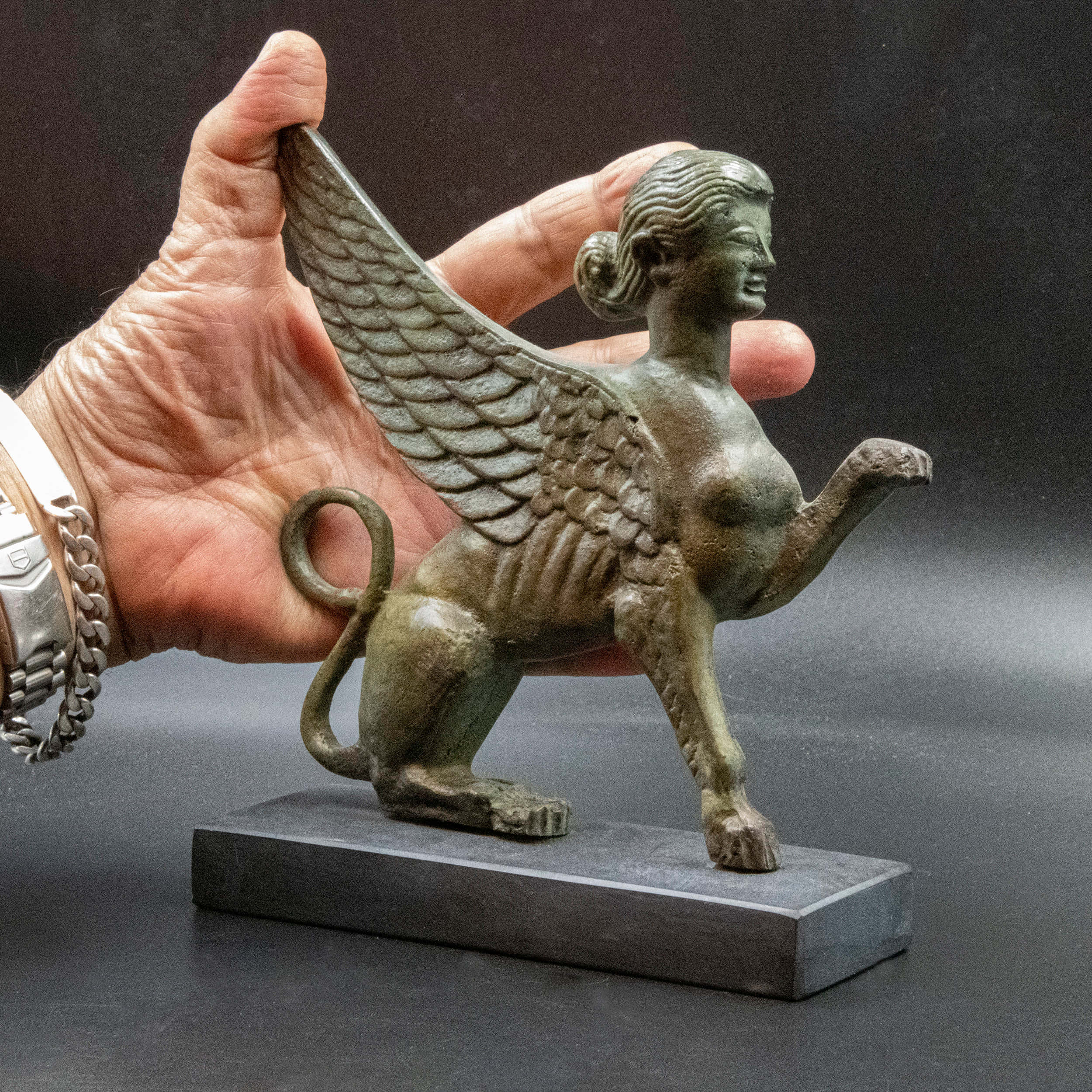 Mystical Artifacts 桌布 壁紙 — 一隻手拿著一個細節豐富的青銅獅身人面像，帶有翅膀和獅身，背景為深色攝影棚。