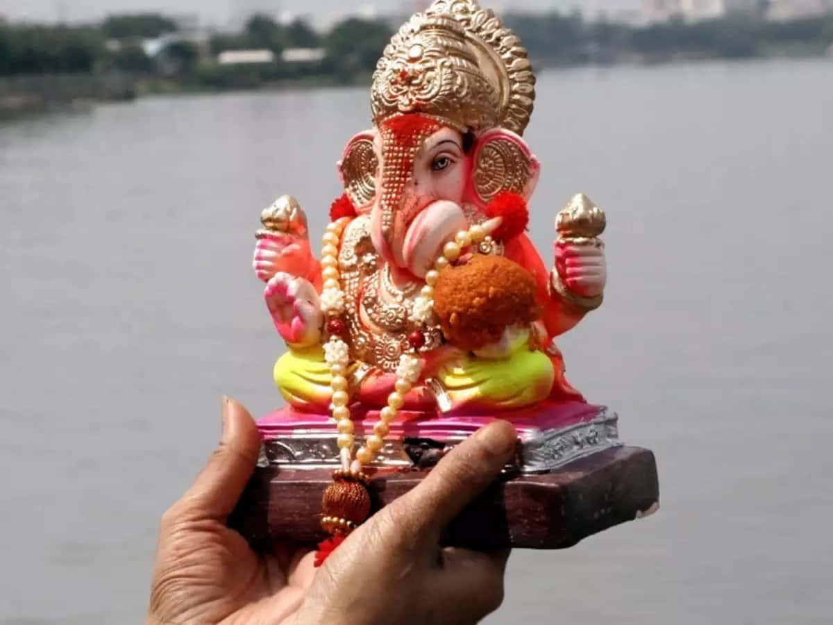 Ganesh Chaturthi 桌布 壁紙 — 一隻手拿著一個色彩繽紛、裝飾華麗的象神像，飾有珠子和黃金，背景是模糊的河流。
