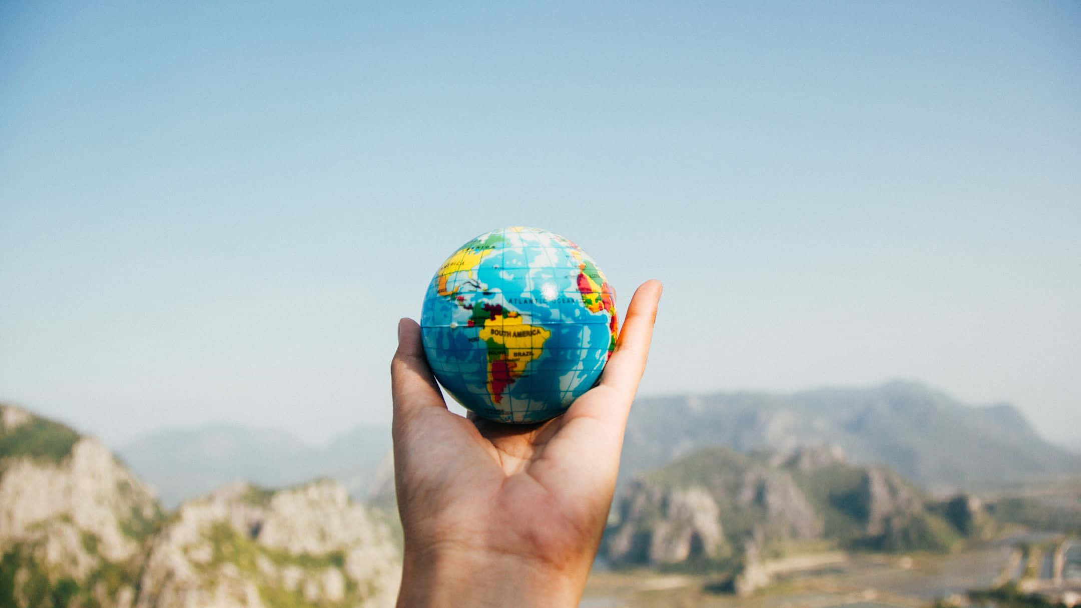 globe 桌布 壁紙 — 一隻手拿著一個色彩繽紛的地球儀，背景是遠處的山脈和晴朗的藍天。
