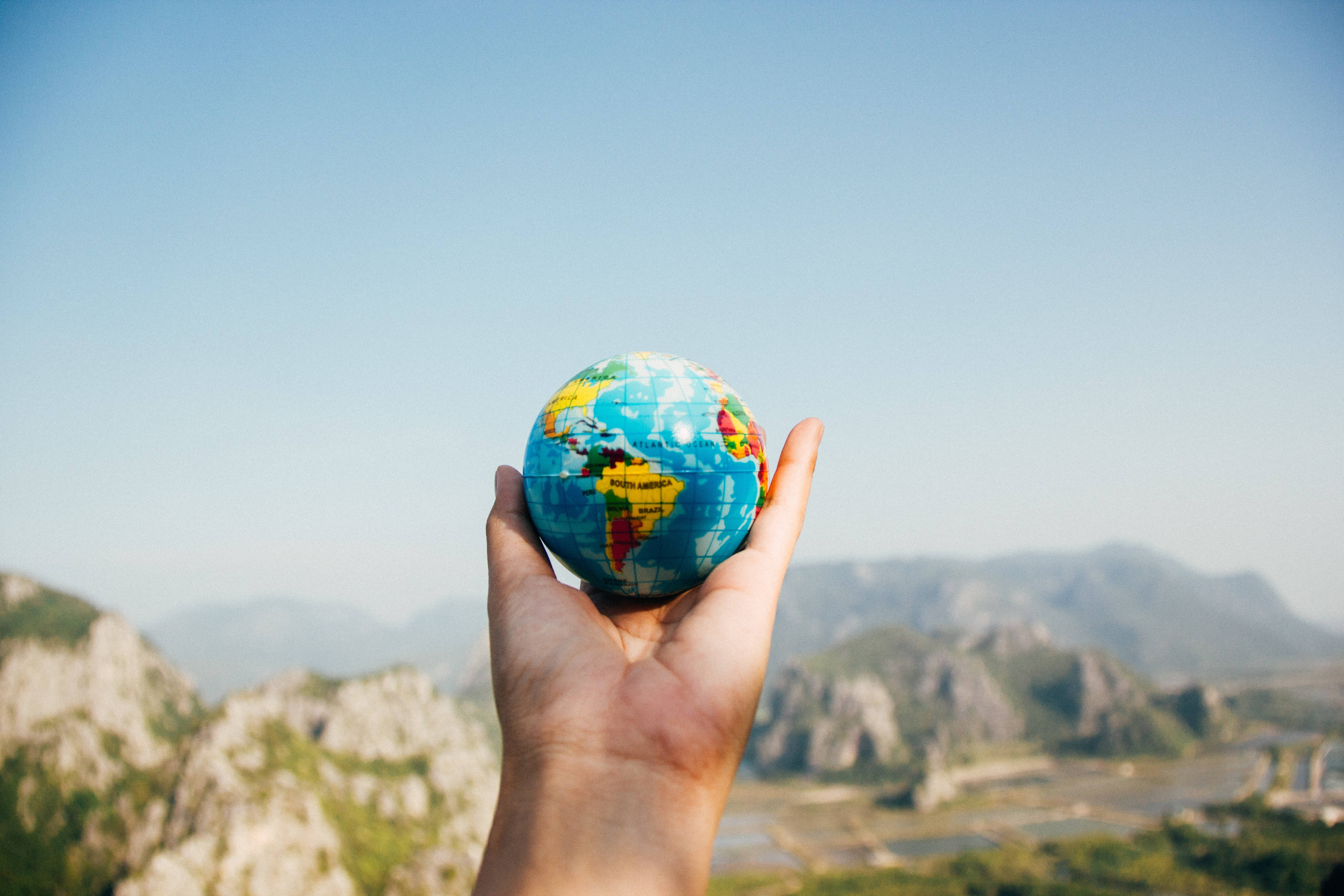 World 桌布 壁紙 — 一隻手拿著一個色彩繽紛的地球儀，背景是遠山和清澈的藍天。