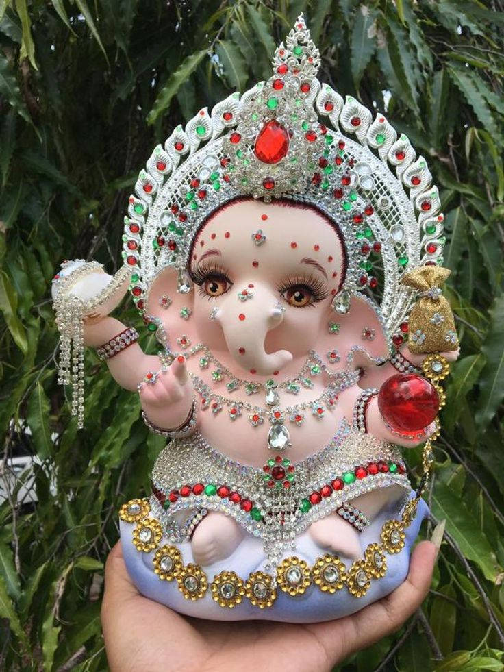laxmi ganesh saraswati 桌布 壁紙 — 一隻手拿著一個華麗鑲嵌寶石的粉紅色象神雕像，飾有紅色、綠色寶石和金色點綴，背景是綠葉。