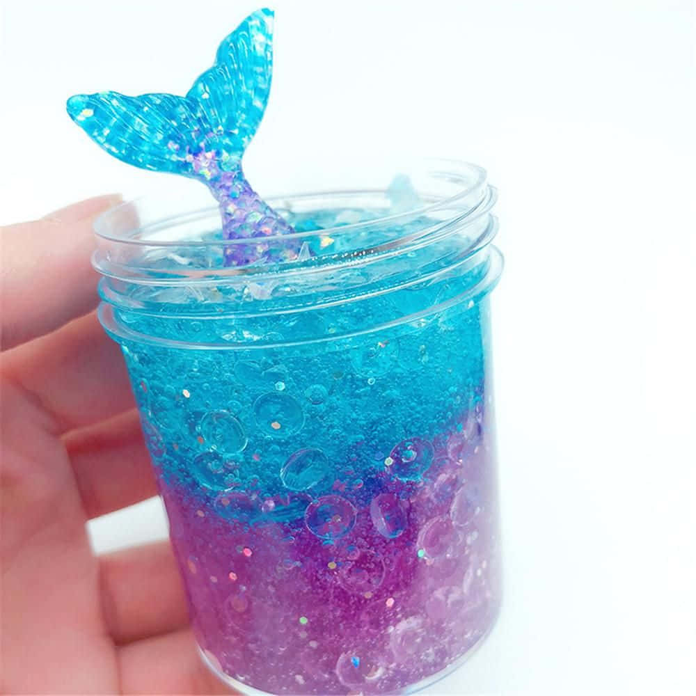 slime 桌布 壁紙 — 一隻手拿著一個裝有閃亮藍色和紫色美人魚史萊姆的罐子，上面有一個閃閃發光的尾巴。
