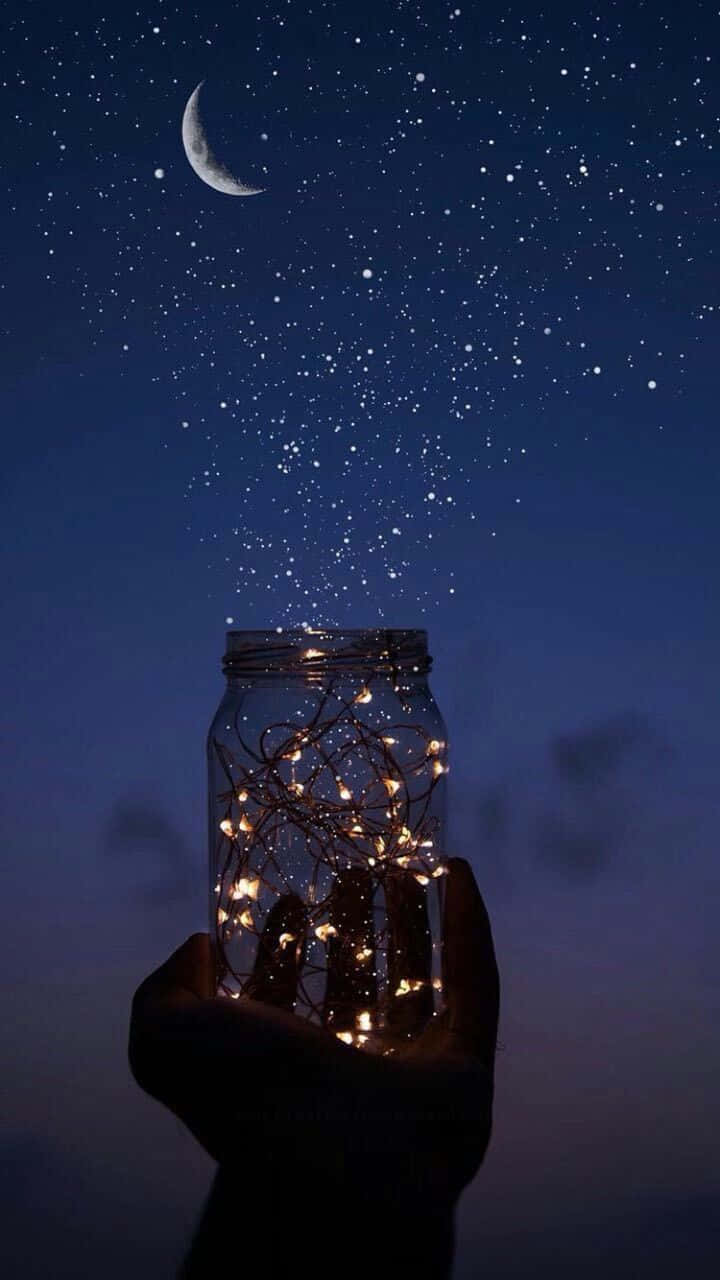 Moon And Stars iPhone 桌布 壁紙 — 一隻手拿著一個裝滿發光仙女燈的罐子，星星和彎月在上方閃爍。