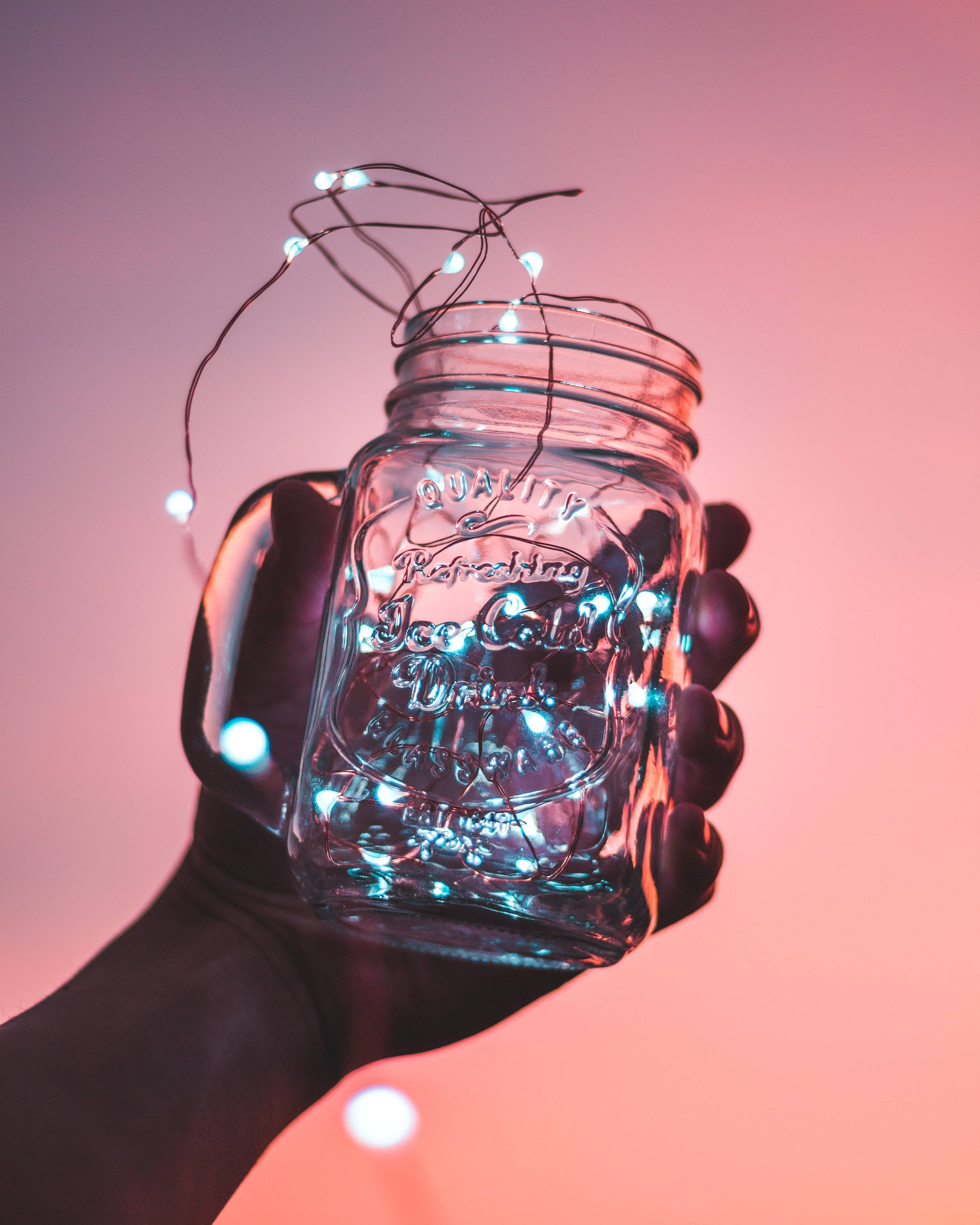 jar 桌布 壁紙 — 一隻手拿著一個裝滿發光燈串的玻璃梅森罐，背景是柔和的粉紅色漸變。