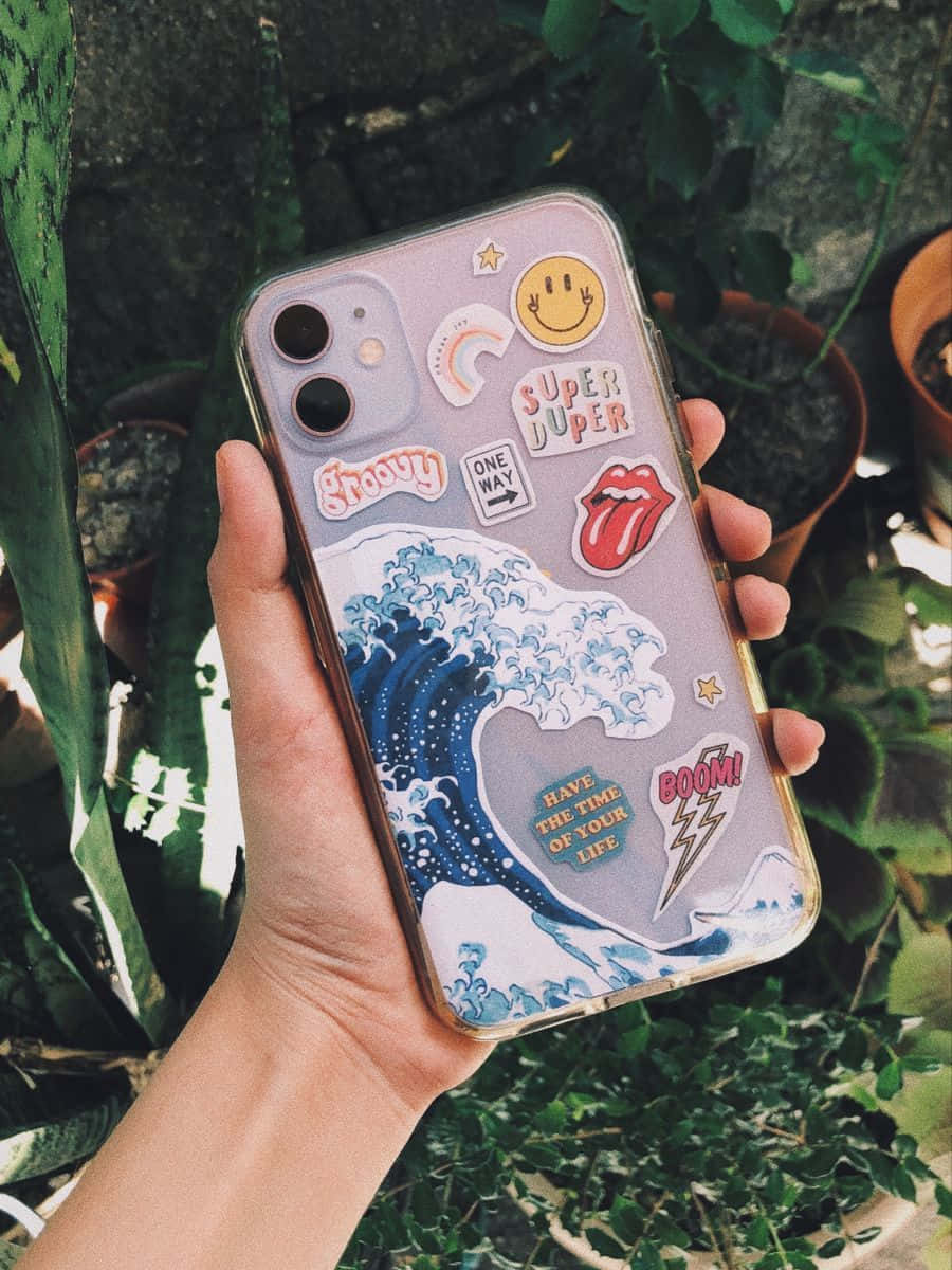 美學 iPhone 壁紙 桌布 壁紙 — 一隻手拿著一個裝飾有彩色貼紙和藍色波浪設計的手機殼，背景是茂盛的綠色盆栽植物。