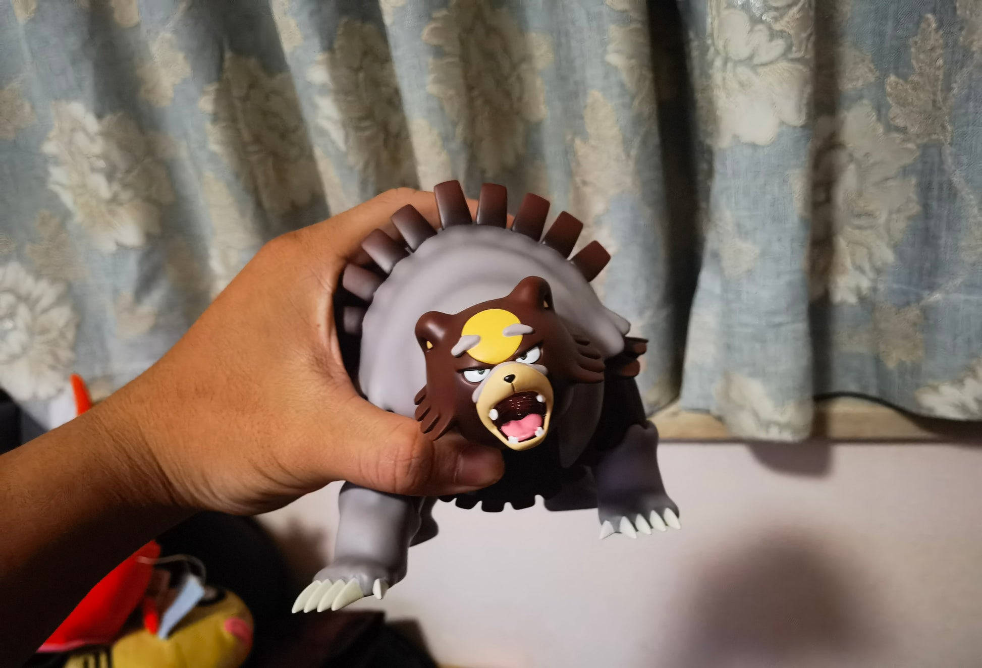 小孩動圖 桌布 壁紙 — 一隻手拿著一個詳細的棕色和灰色Ursaring Pokémon公仔，背部有尖刺，表情憤怒，背景是花卉窗簾。