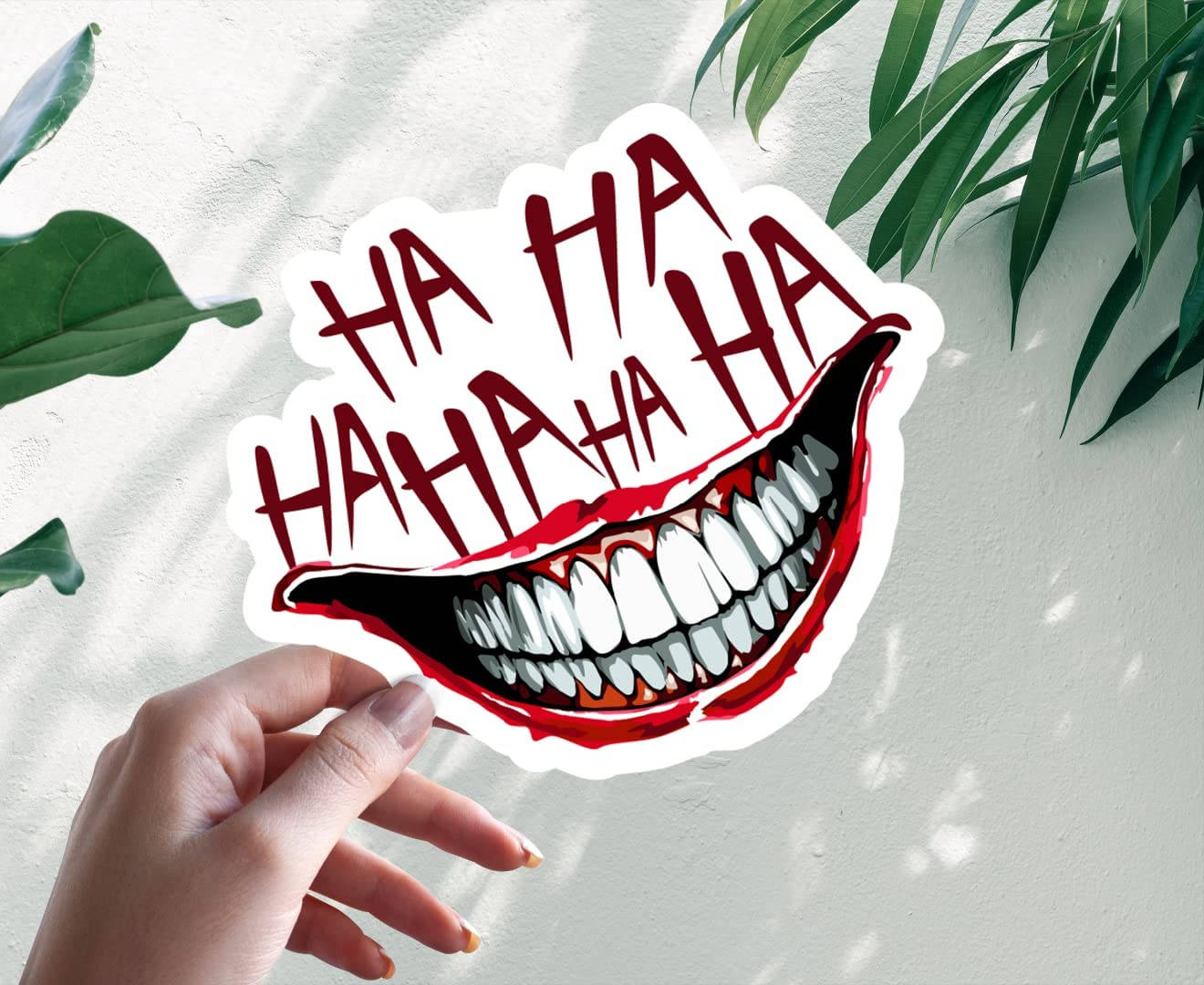 Hahaha 桌布 壁紙 — 一隻手拿著一個貼紙，上面有張開大笑的嘴巴，露出尖牙，紅色「HA HA HA」文字，背景是白色牆壁和綠葉。