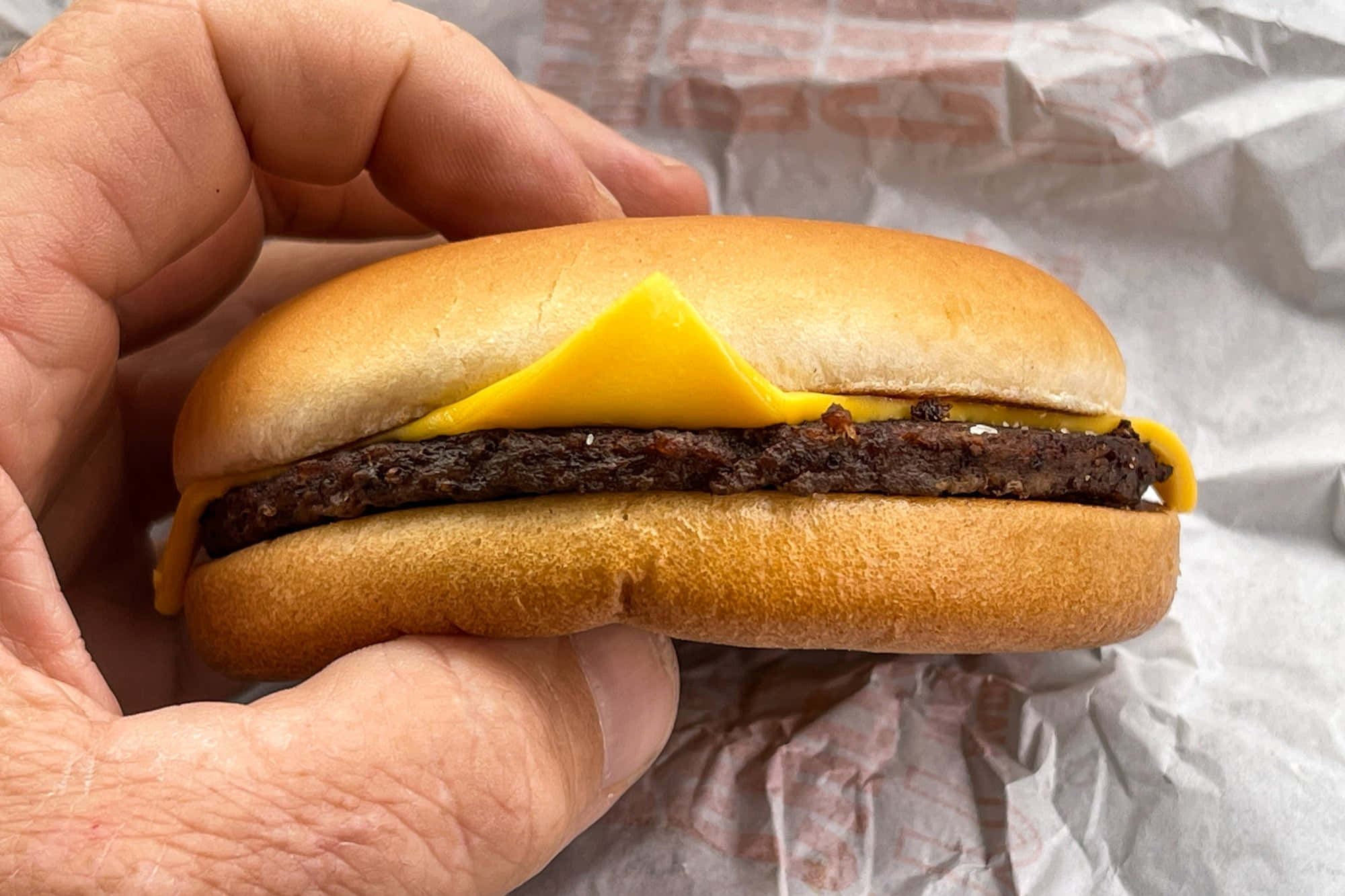 fast food 桌布 壁紙 — 一隻手拿著一個起司漢堡，融化的黃色起司片從烤麵包和多汁牛肉餅之間露出來。