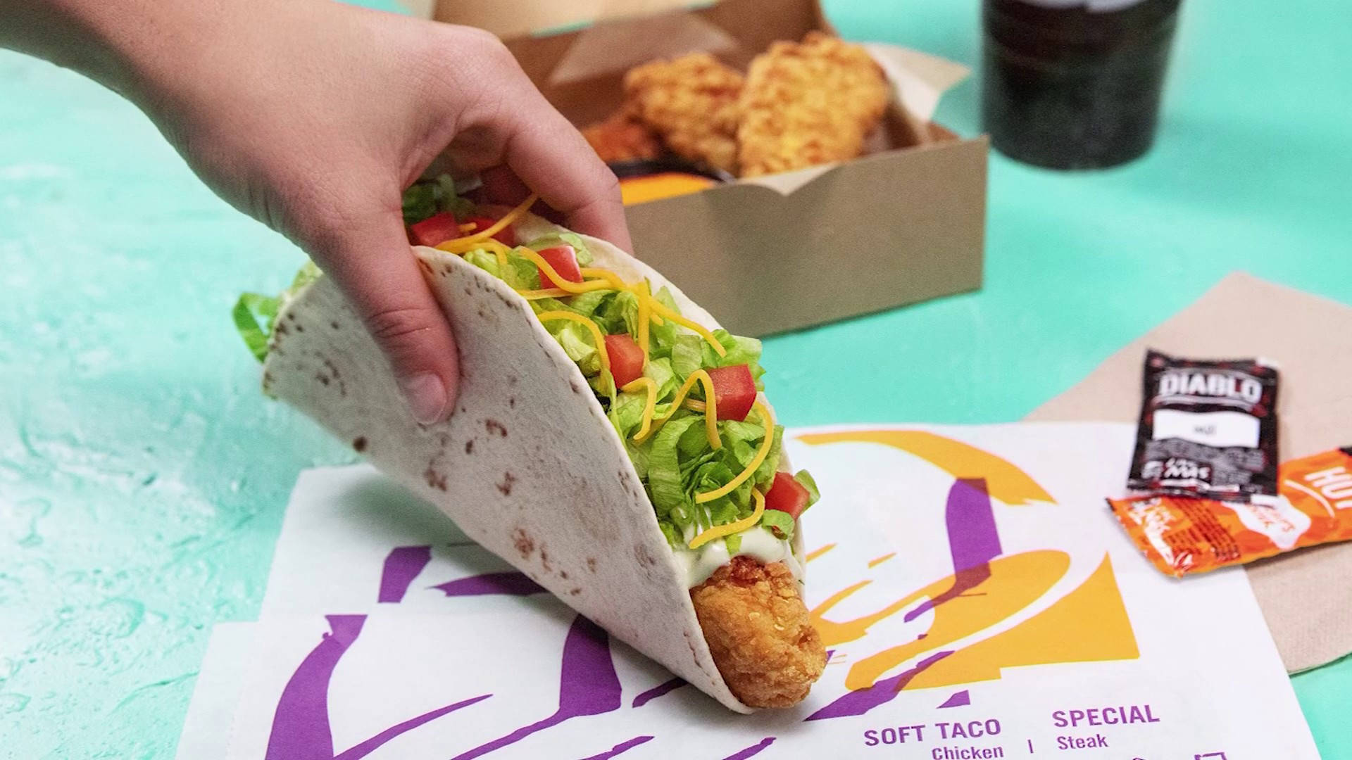 Taco Bell 桌布 壁紙 — 一隻手拿著一個軟玉米餅，裡面有脆雞肉、生菜、起司和番茄，放在有品牌標誌的紙上，背景是青綠色。