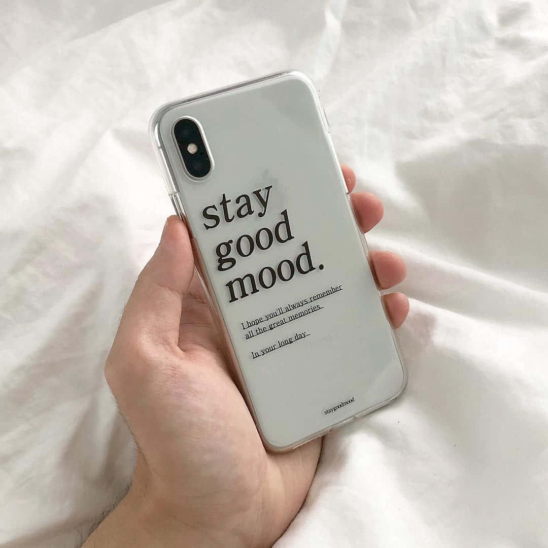 iPhone 美學 桌布 壁紙 — 一隻手拿著一個透明手機殼，上面有 'stay good mood' 字樣，背景是柔軟的白色床單。