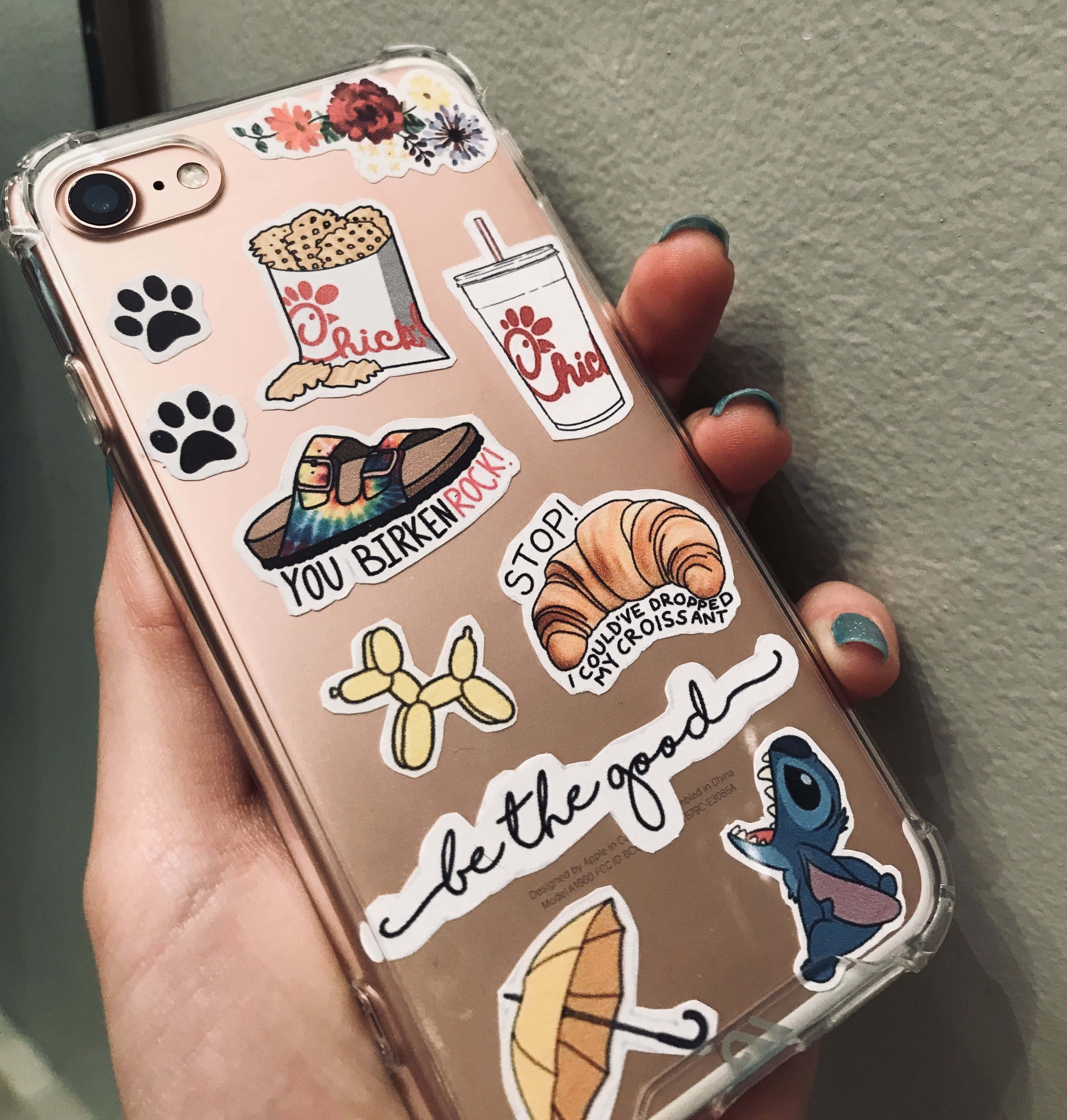 Phone Case 桌布 壁紙 — 一隻手拿著一個透明手機殼，上面貼滿了彩色貼紙，包括Chick-fil-A食物、腳印、Birkenstock鞋、可頌麵包、史迪奇和勵志語句。