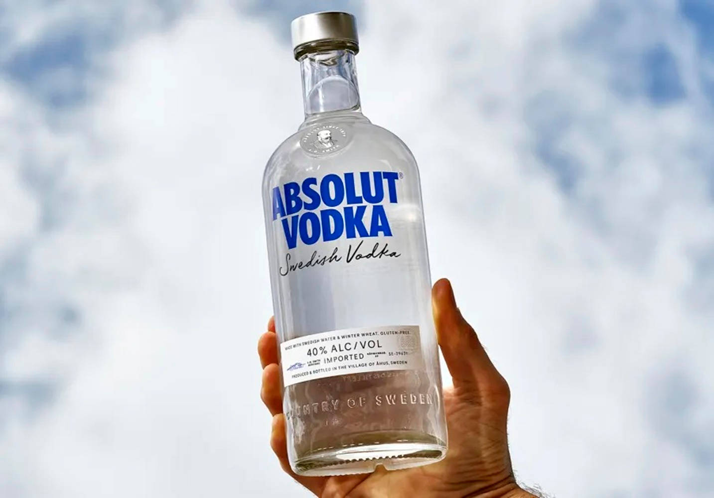 bottle 桌布 壁紙 — 一隻手拿著一個透明的Absolut Vodka酒瓶，背景是明亮的陰天天空。