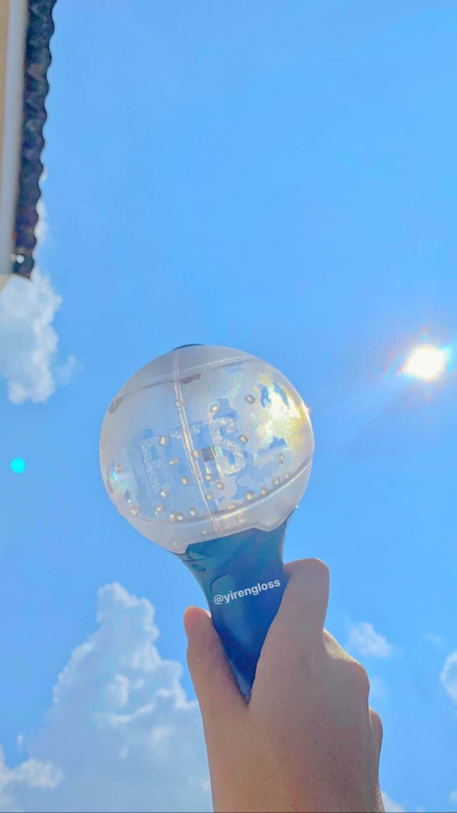 Bts Army Bomb 桌布 壁紙 — 一隻手拿著一個透明閃亮發光的手機殼，背景是晴朗的藍天、蓬鬆的雲朵和太陽光暈。