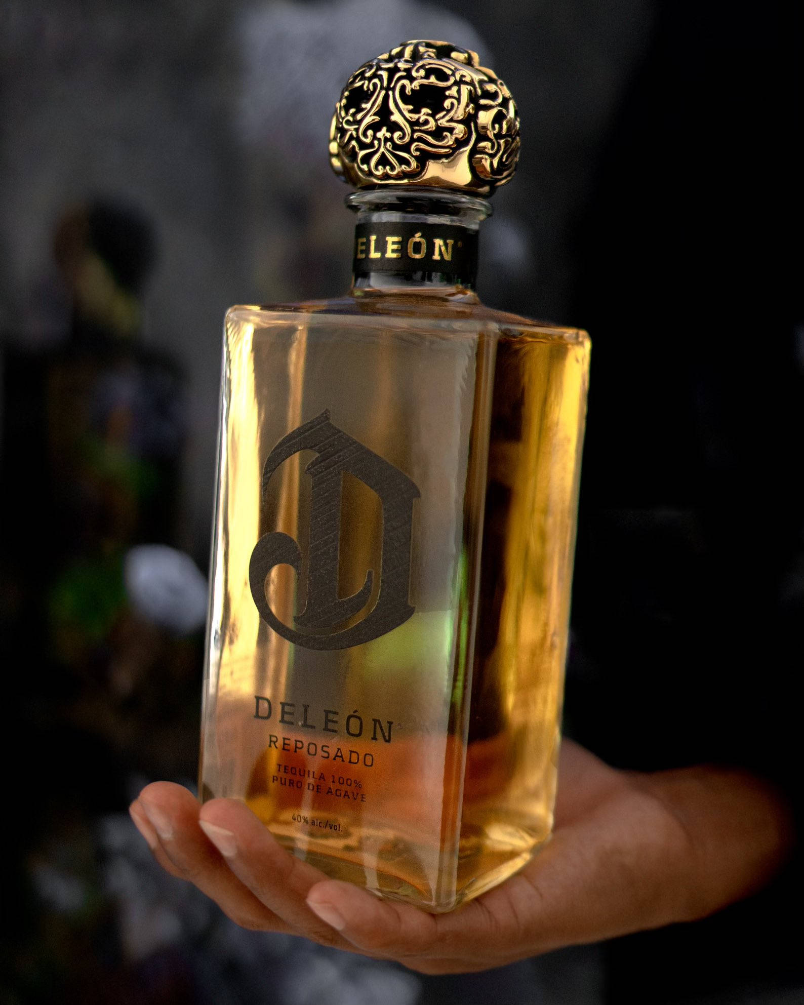bottle 桌布 壁紙 — 一隻手拿著一個金色的Deleón Reposado龍舌蘭酒瓶，帶有華麗的金色瓶蓋，背景模糊黑暗。