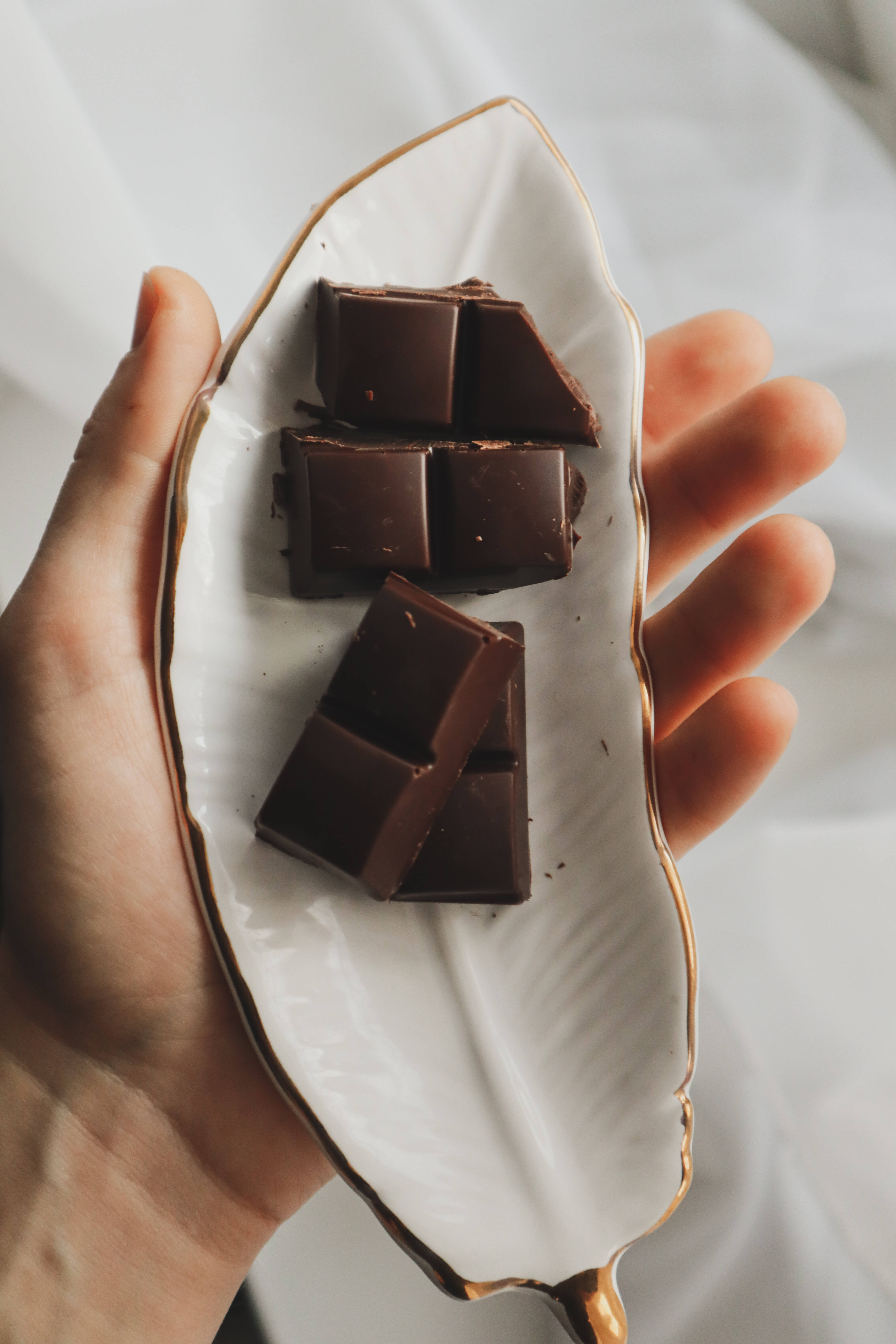 Chocolate 桌布 壁紙 — 一隻手拿著一個金色邊緣的葉形白色盤子，在柔軟的白色布料上展示濃郁的黑巧克力方塊。