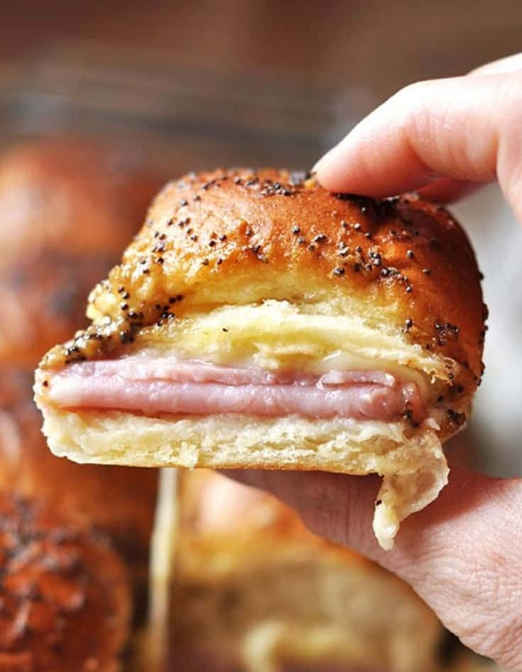 Black Forest Ham 桌布 壁紙 — 一隻手拿著一個金黃色的罌粟籽火腿起司小漢堡，露出酥脆的層次和融化的起司。