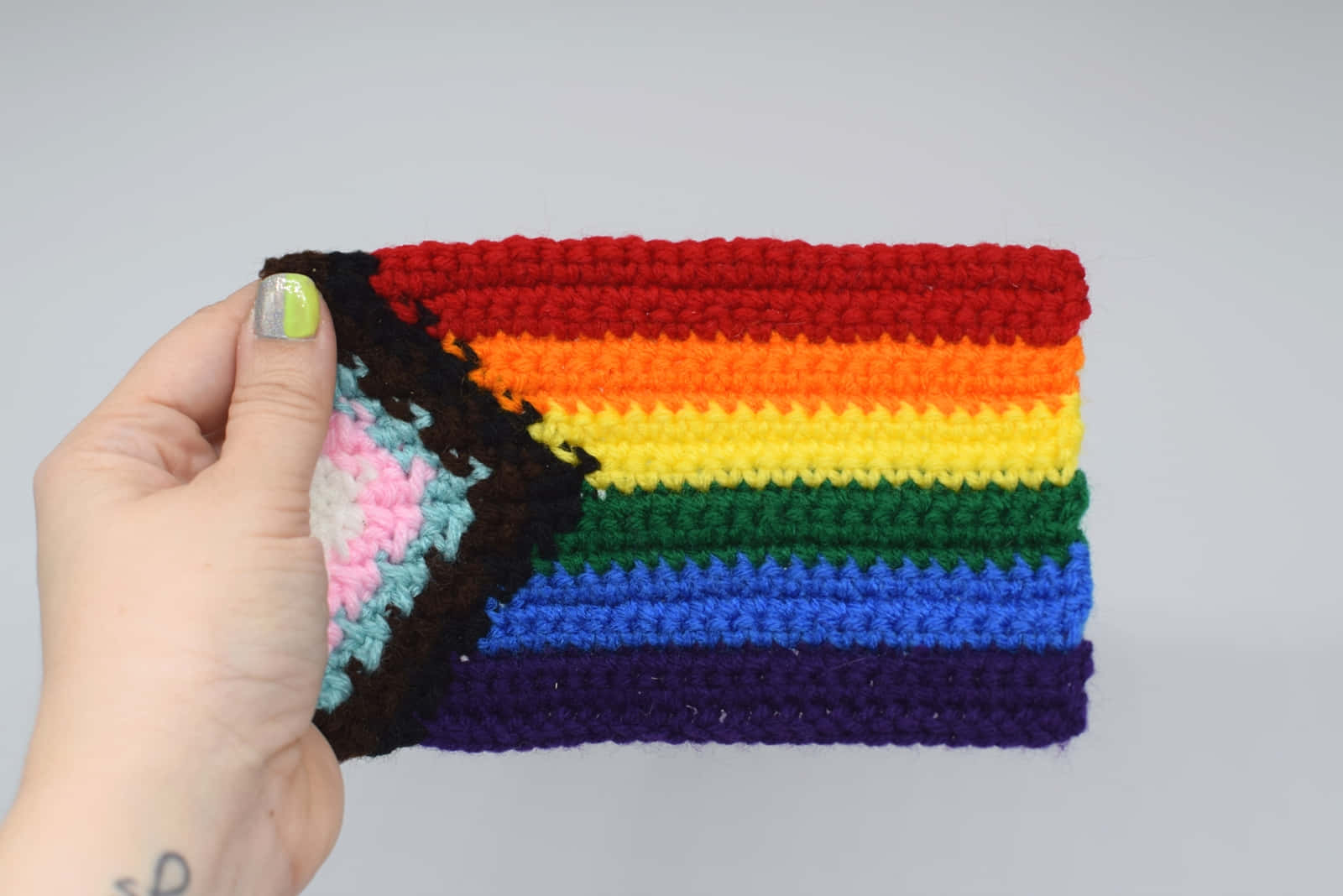 Pride Flag 桌布 壁紙 — 一隻手拿著一個鉤織的彩虹驕傲旗幟，帶有棕色邊框，背景為純灰色。