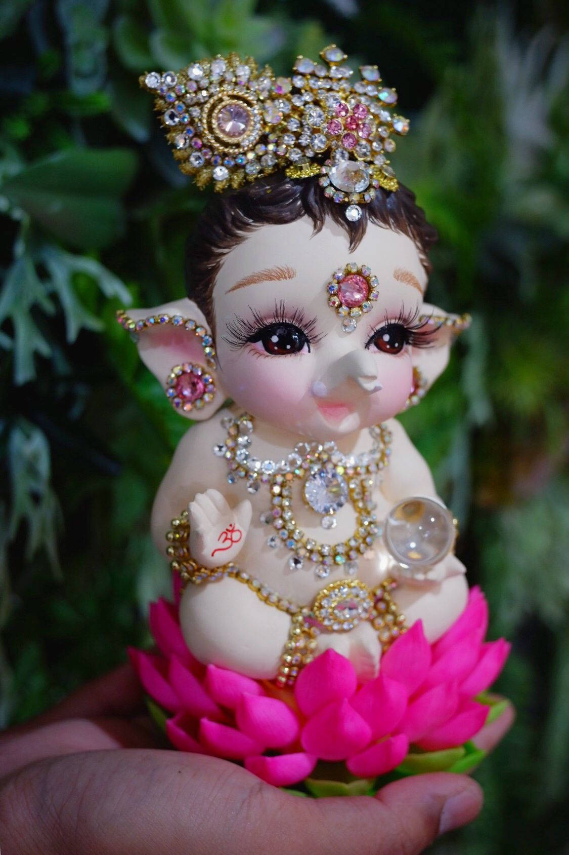 cute ganesha 桌布 壁紙 — 一隻手拿著一個閃閃發光的象神雕像，飾有珠寶，坐在明亮的粉紅色蓮花上，背景是綠色樹葉。