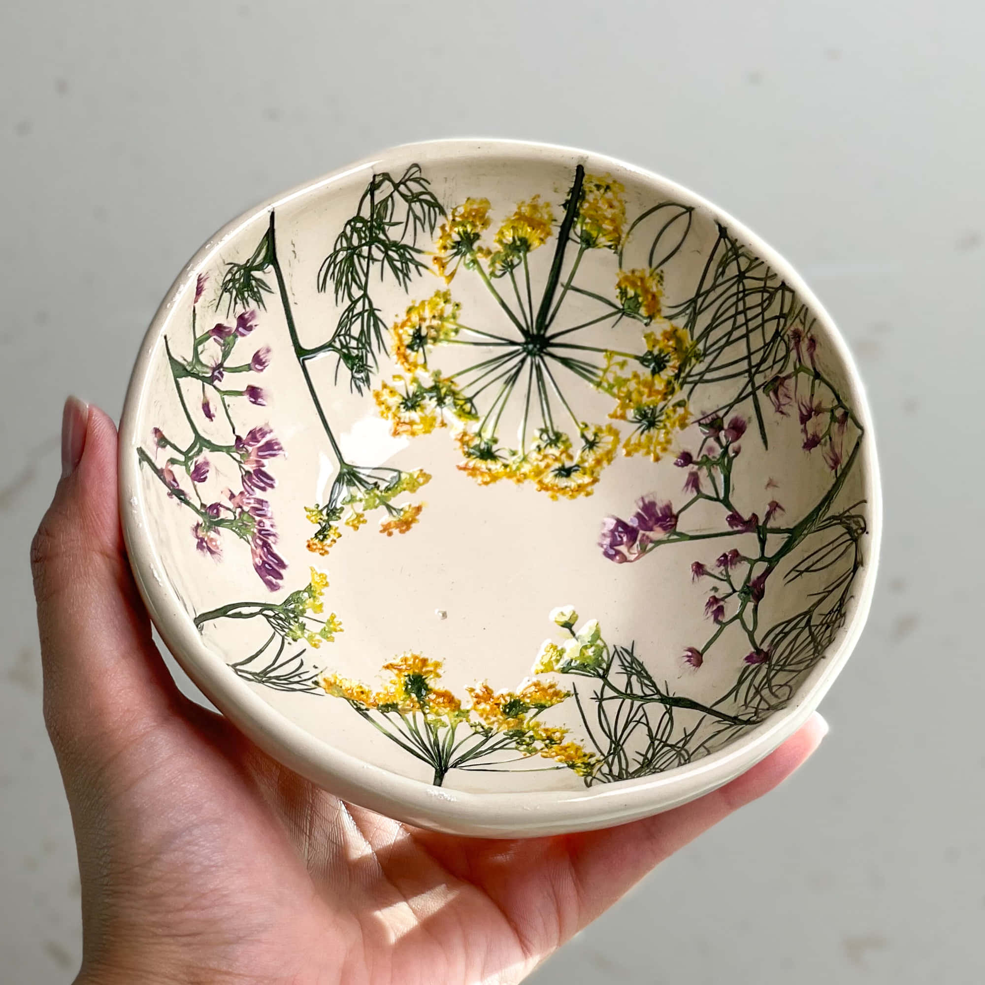 Ceramic 桌布 壁紙 — 一隻手拿著一個陶瓷碗，上面裝飾著壓製的黃色和紫色野花，背景是柔和的白色。