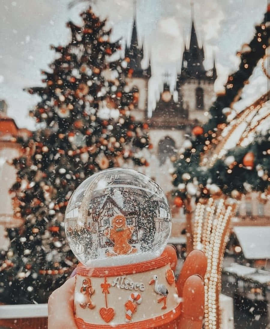 聖誕美學 桌布 壁紙 — 一隻手拿著一個雪球，裡面有一個薑餅人，背景是白雪覆蓋的聖誕樹和城堡。