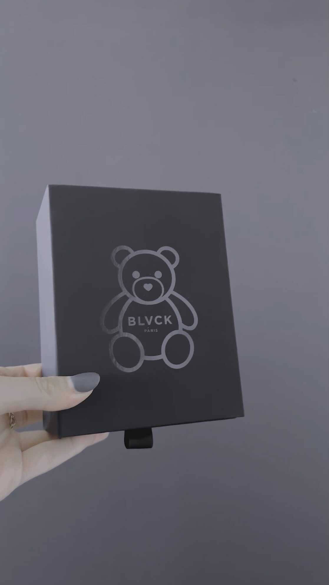 Blvck Paris 桌布 壁紙 — 一隻手拿著一個霧面黑色盒子，上面有簡約的銀色泰迪熊標誌和 BLVCK PARIS 文字，背景為純灰色。